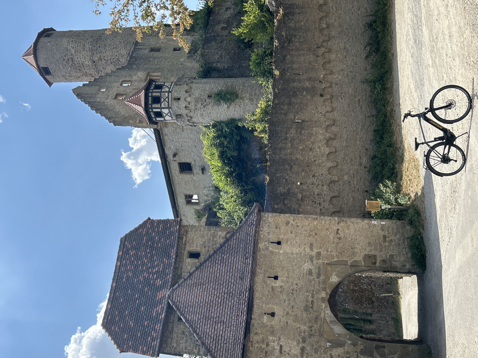 A sunlit, full shot captures the historic German castle, Burg Hornberg, in Neckarzimmern. The castle, perched on a hillside, features a formidable stone wall, an arched gateway, and a prominent round tower with a red-tiled conical roof. An upper section of the castle reveals weathered stone walls and battlements, with a smaller turret adorned with a half-timbered upper section and a red-tiled roof. Lush green vegetation grows along the castle walls and slopes. In the foreground, a modern black electric bicycle stands on a gravel path, its kickstand deployed. The sky above is a vibrant blue, dotted with fluffy white clouds.