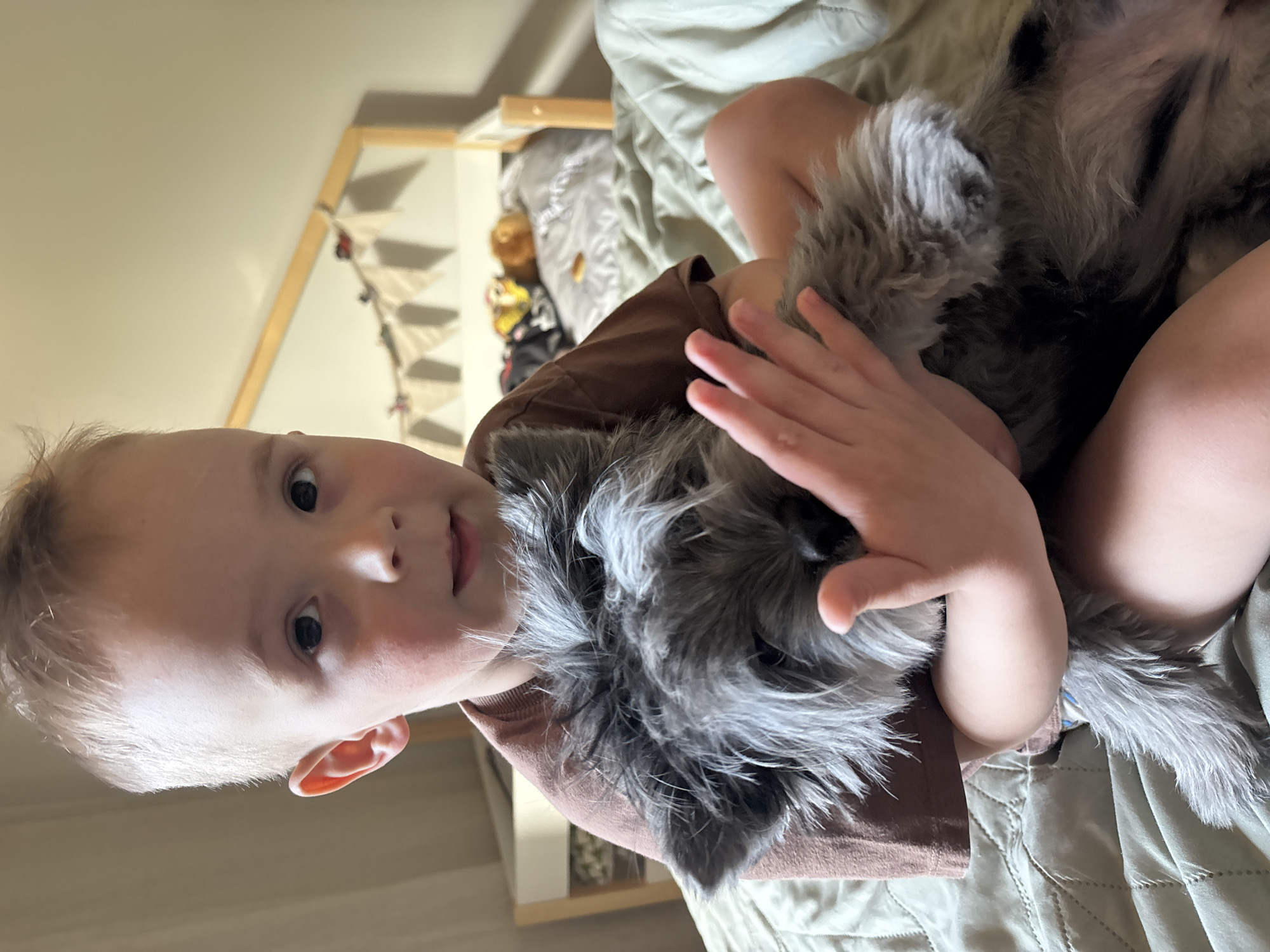 A young child with blond hair and fair skin is sitting on a bed covered with a light green quilted bedspread. The child is wearing a brown t-shirt and is holding a gray, furry toy dog. The child's right hand is placed on the toy's head. Behind the child, a wooden bed frame is visible, decorated with small white pennant flags. The location is the city of Chaikovsky, Russia.