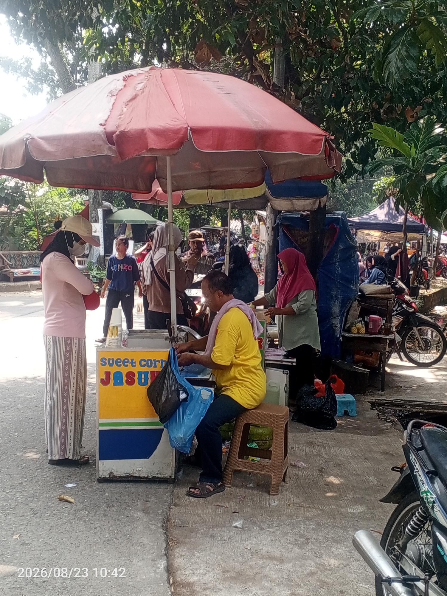 A street scene in Depok, Indonesia, features a food stall with a large red umbrella. In the foreground, a man in a yellow shirt is preparing food, with a blue plastic bag and a black bag beside him. A woman in a pink shirt and a patterned skirt stands to the left, wearing a face mask and hat. Behind them, several other people are visible, some standing and some sitting. The stall itself is a yellow cart with "sweet corn JASUK" written on it, and a green umbrella can be seen in the background. The environment is outdoors, with trees and foliage providing shade. Several motorcycles are parked nearby, and other stalls are set up in the distance, suggesting a market or roadside vendors. The timestamp on the image reads "2026/08/23 10:42".