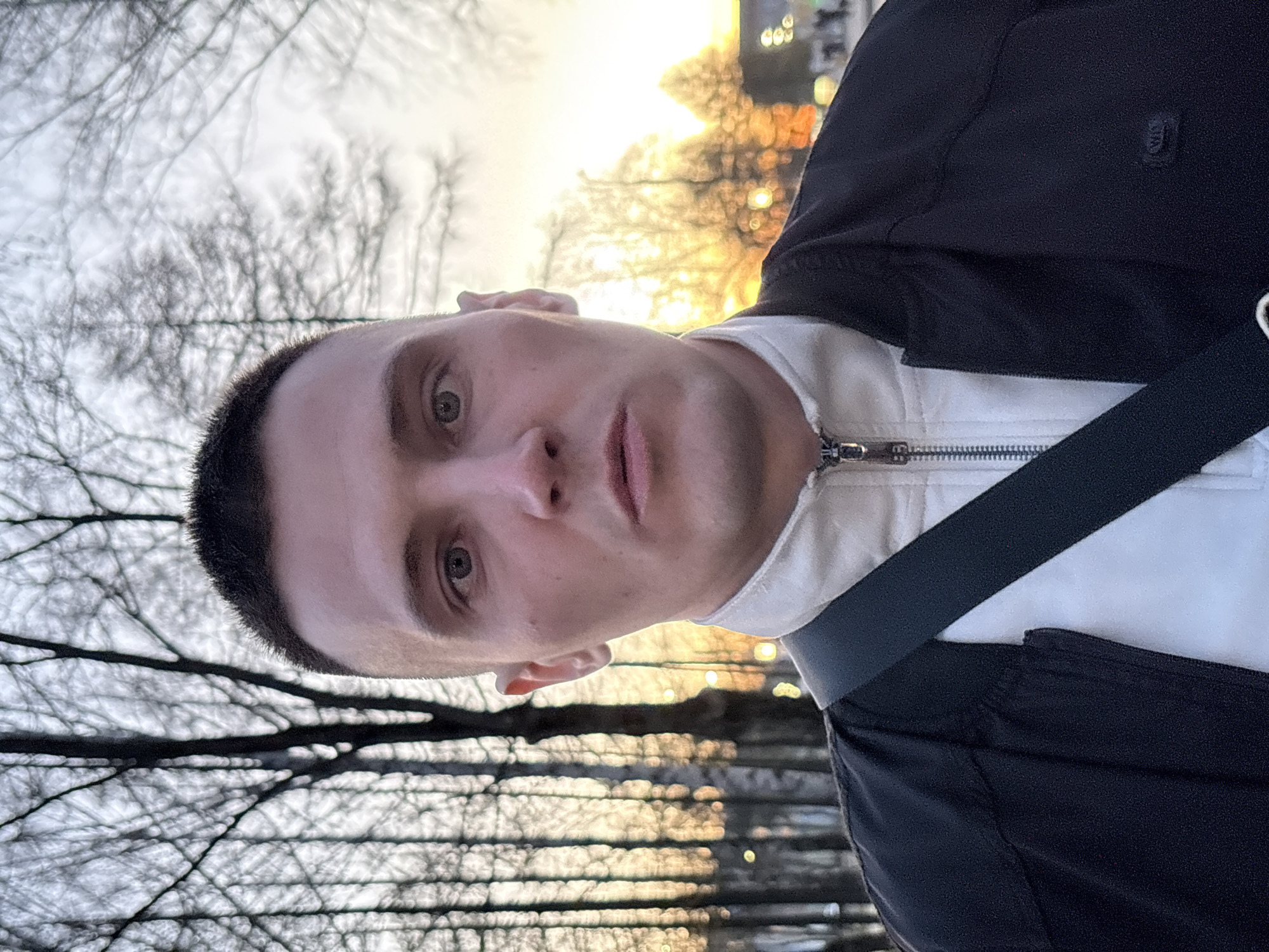 A man is taking a selfie outdoors. He has short, dark hair and light eyes, and he is wearing a white jacket with a black jacket over it. A black strap is across his chest. The background shows bare trees and what appears to be a body of water reflecting light. The location is Chaikovsky, Russia. The lighting suggests either sunset or sunrise.