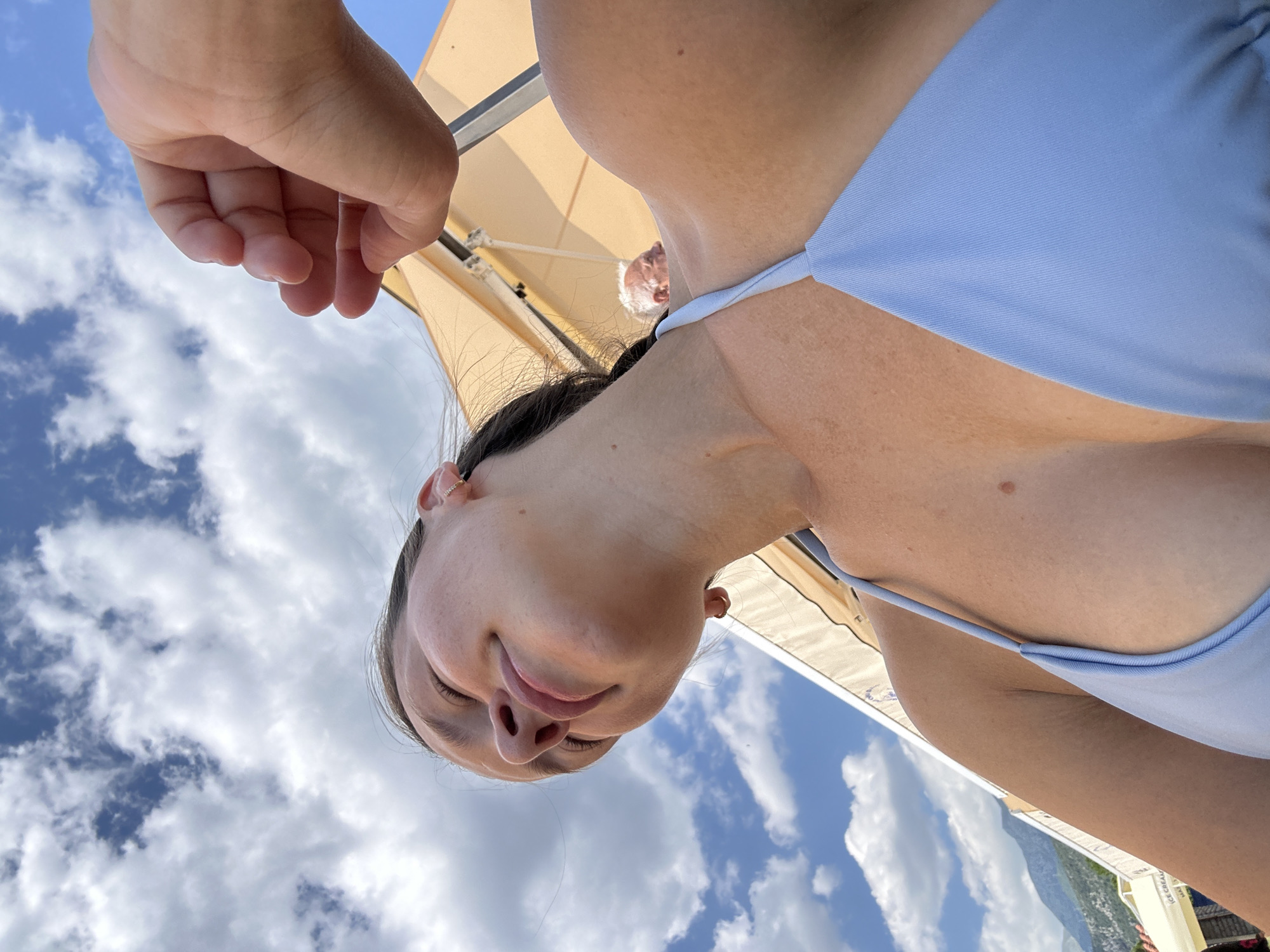 A low-angle, close-up shot captures a young woman from the chest up, wearing a light blue bikini top. Her eyes are closed, and her head is tilted back, suggesting relaxation. A hand is visible in the upper right portion of the frame, reaching towards her face. The background features a bright blue sky filled with fluffy white clouds, and hints of a beige awning and distant mountains, indicative of a coastal resort setting. The image appears to have been taken outdoors on a sunny day, likely in a location like Budva, Montenegro, known for its beaches and scenic beauty. An older man is partially visible in the background, out of focus, under the awning.