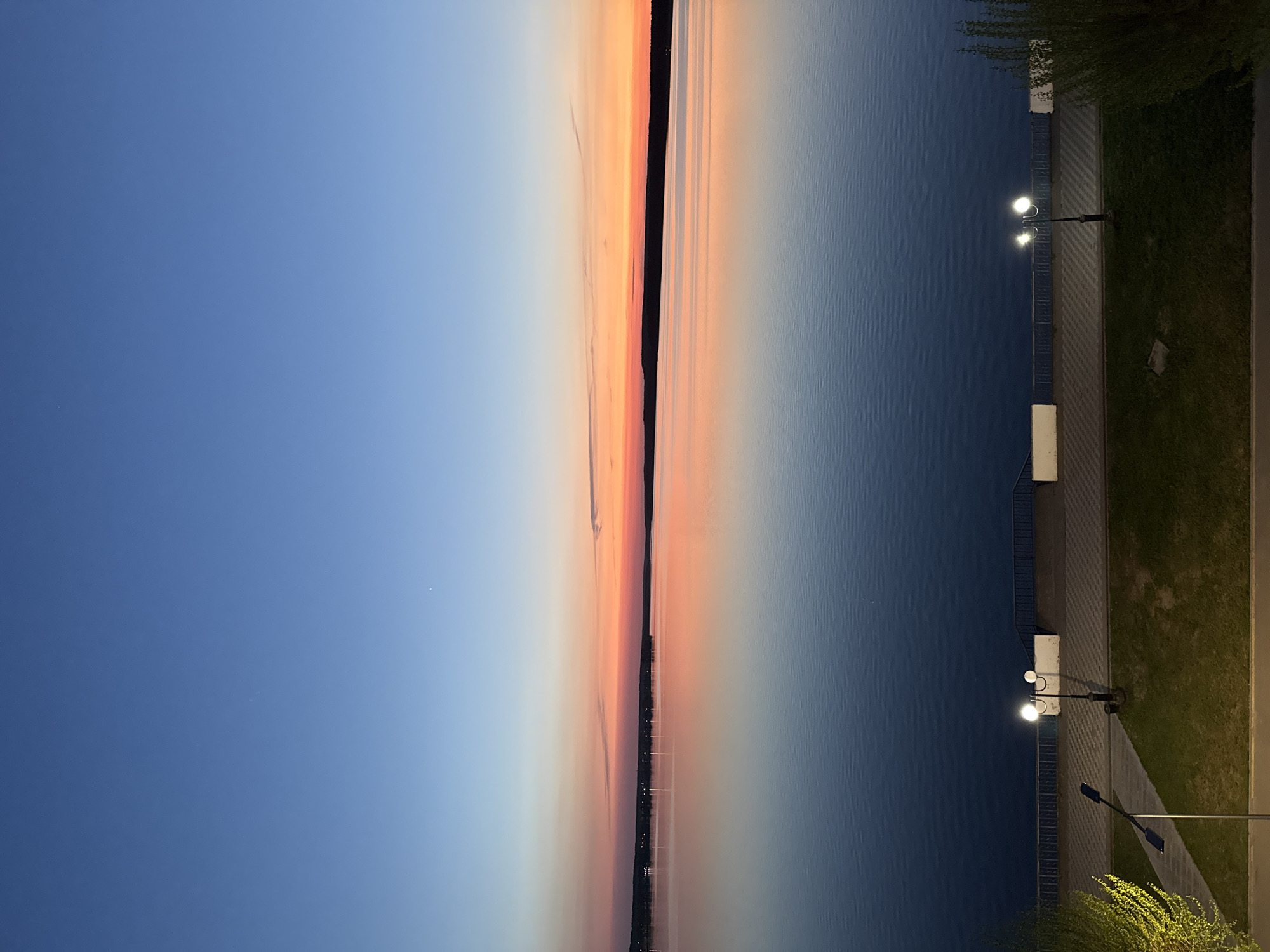 The image presents an evening view in Votkinsk, Russia. The scene is bisected vertically, showing a transition from a clear blue sky to a body of water, likely the Votkinsk Reservoir. A thin strip of land is visible between the sky and the water, marked by a sunset gradient of orange and pink. On the right side, bordering the water, is a concrete embankment with streetlights illuminating a paved path alongside a grassy area. The lights indicate it is after sunset. The overall impression is that of a calm, tranquil evening atmosphere.