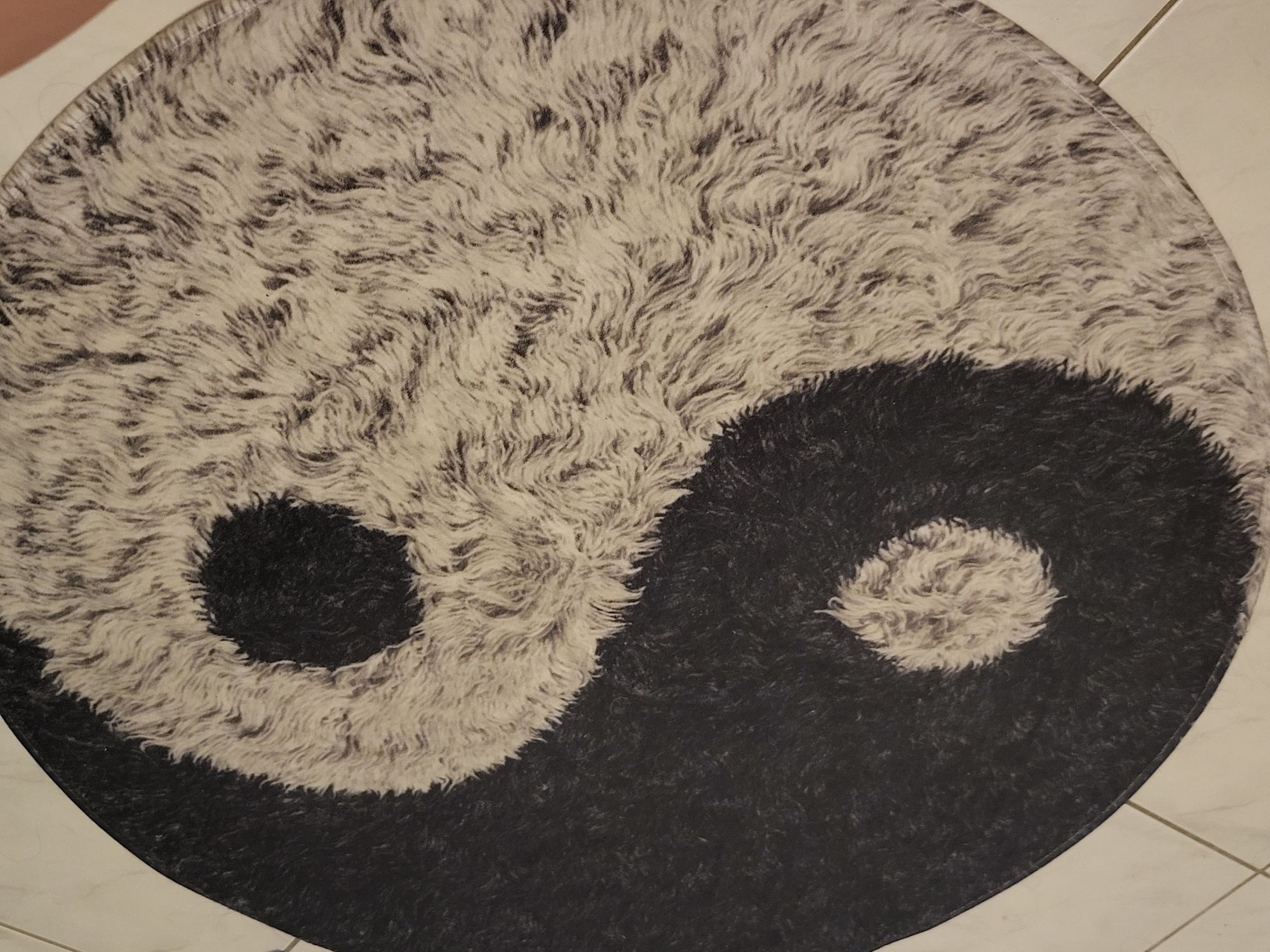 The image shows a close-up, overhead view of a round, plush rug designed with a Yin and Yang symbol. The rug is on a tiled floor with light-colored square tiles and grout lines visible at the edges of the image. The Yin and Yang symbol is depicted with a black, textured half and a lighter, cream-colored textured half, both featuring small circular inlays of the opposite color. The rug's texture is dense and appears to be made of short, wavy fibers, giving it a furry appearance. The lighting is even, and there are no people or distinct environmental features within the rug's depiction. The image does not provide any information about its specific location in Pinneberg, Germany, beyond it being placed on a floor.