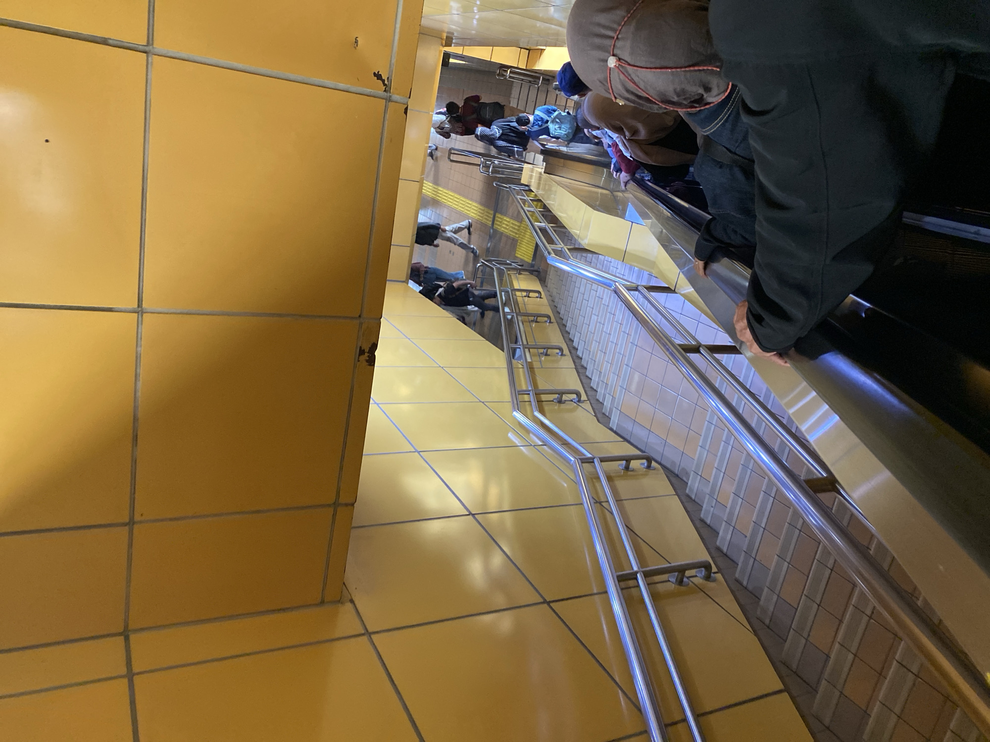 The image depicts an underground passage or walkway with yellow tiled walls and ceiling. Multiple people are present, some descending on an escalator and others walking on a tiled floor. There are also staircases with stainless steel railings visible. The scene appears to be a public transportation hub or a connecting passage within a building. The lighting suggests it is indoors. The location is identified as Central Jakarta, Indonesia.