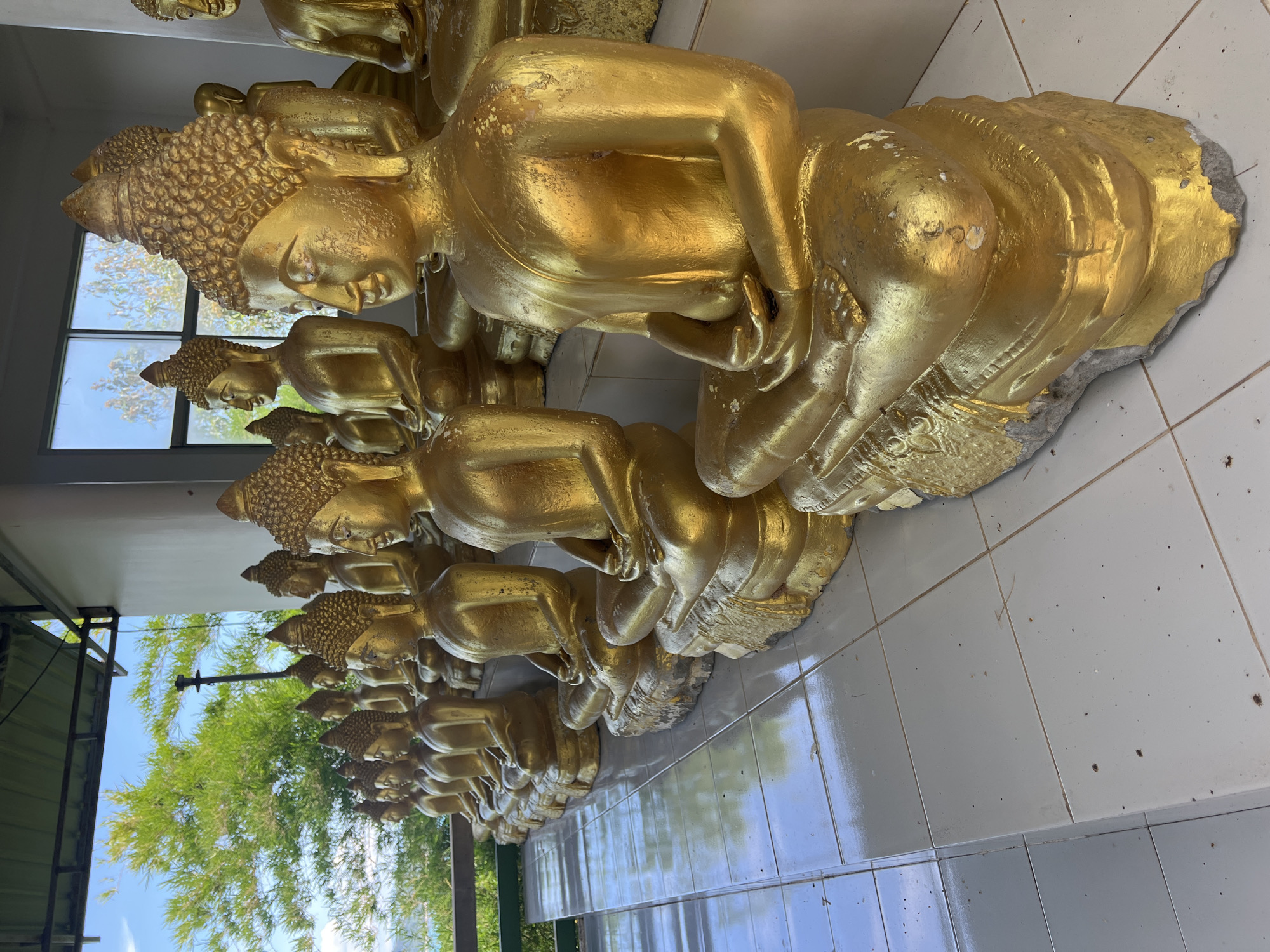 The image depicts a row of golden Buddha statues sitting in meditation. The statues are arranged on a white tiled floor, under a covered walkway with a metal roof. In the background, lush green trees and a bright blue sky with scattered clouds are visible. The statues are made of a material that appears to be painted gold, with some wear and tear visible on the surface of the statues, particularly on their bases. The overall scene suggests a tranquil and spiritual setting, possibly a temple or a place of worship in Baan Thong Krut, Thailand, given the architectural style and the presence of Buddhist iconography.