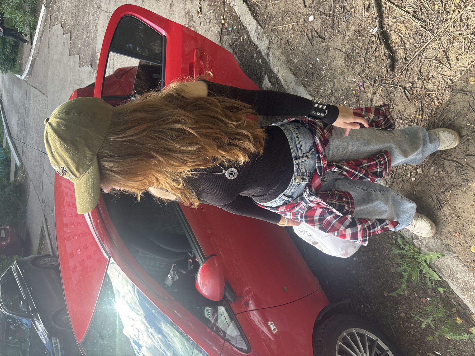 A young person with long, blonde hair, wearing a green baseball cap, a black long-sleeved shirt, and jeans, is leaning against a red car with the driver's side door open. The person has a red and black plaid shirt tied around their waist, and a pentagram necklace. They are holding a white plastic bag in their right hand. The scene appears to be outdoors, possibly a street or a parking lot, with a rough, dirt-covered ground and some vegetation. The lighting suggests it is daytime, and the weather seems to be fair. The overall mood is casual, and the person seems relaxed, possibly having just finished shopping or is about to go somewhere. There are no visible text elements in the image. The location context is Chaikovsky, Russia.