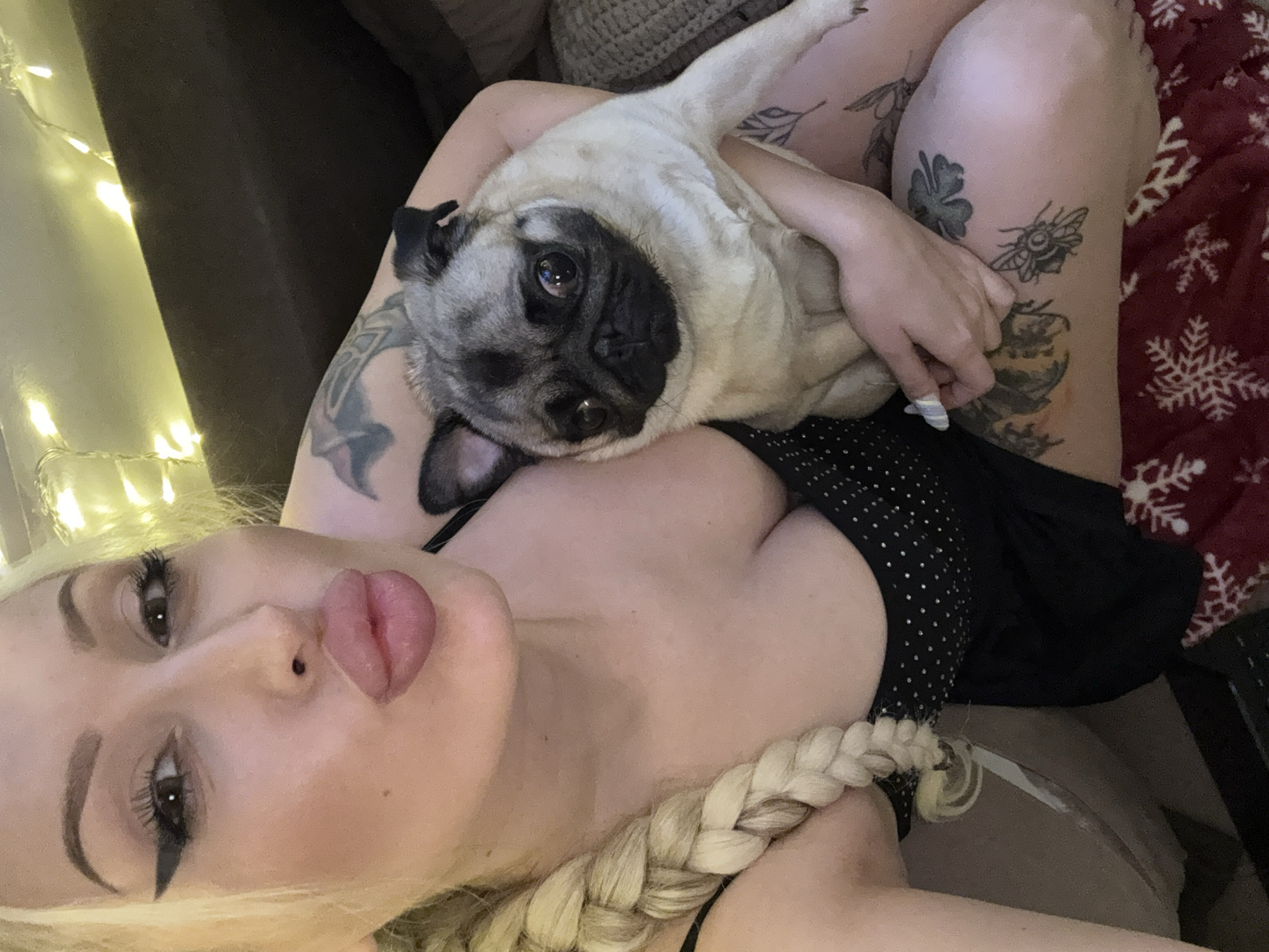 In a dimly lit indoor setting, a woman with blonde hair in a braid and heavy makeup is holding a pug. The woman is wearing a black top with white polka dots, and has visible tattoos on her arms and legs. The pug is light brown and cream-colored. Fairy lights are visible in the background.