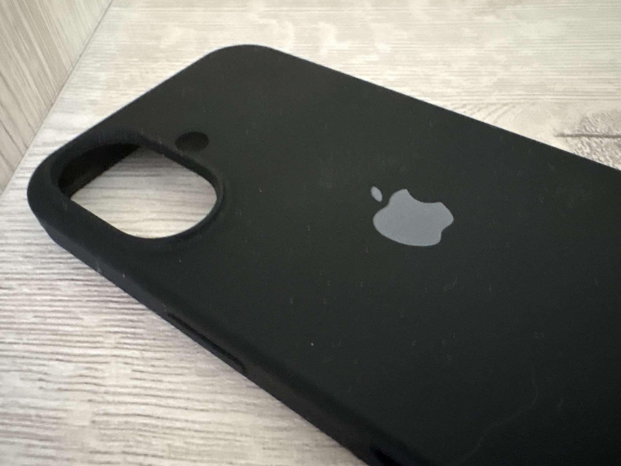 A black iPhone case with a gray Apple logo is placed on a light-colored, weathered wooden surface. The case is positioned at an angle, with the bottom edge closer to the viewer. A circular cutout, likely for a camera lens, is visible on the top left of the case. The wooden surface has a visible grain pattern, suggesting it might be a table or floor. The lighting appears to be natural and somewhat dim, casting subtle shadows. There is no text visible in the image. The scene is static, with the object at rest. The setting is likely indoors, given the lighting and the nature of the object.