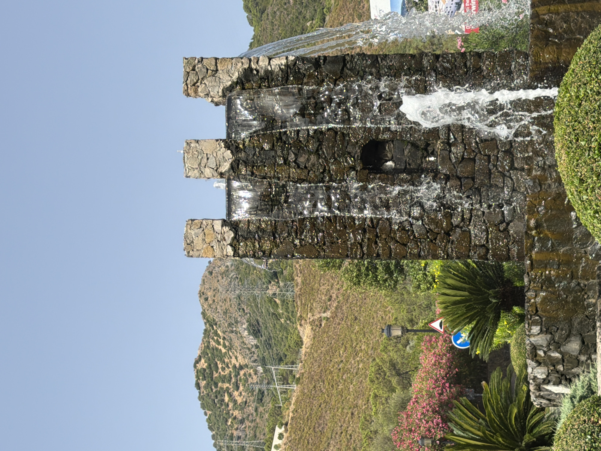This image depicts an outdoor scene featuring a stone fountain with water cascading down its facade. The fountain is constructed from rough-hewn stones and has a crenellated top, resembling a miniature castle tower. Water flows from openings at the top and creates a dynamic, sparkling effect. In the foreground, lush green bushes and plants surround the base of the fountain. Behind the fountain, a street lamp and road signs are visible, suggesting an urban or semi-urban environment. Further in the background, a hillside covered in vegetation rises, with power lines and transmission towers present, indicative of the mountainous terrain surrounding the area. The sky is clear and blue. The scene is likely located in Benahavís, Spain, a town known for its hilly landscape and traditional architecture.