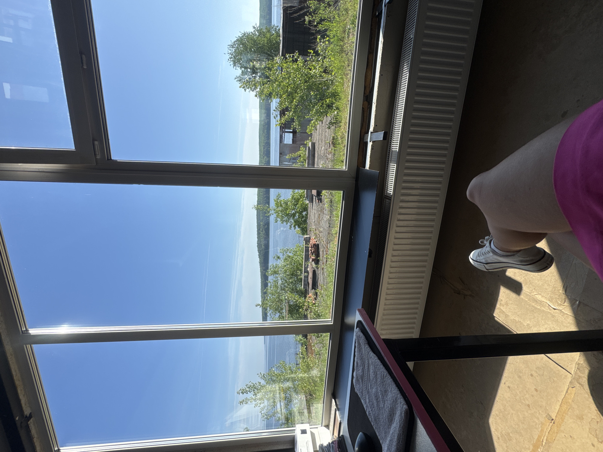 The image shows an interior view through a window with a lake visible outside. The lake appears to be lined with trees and some structures. The sky is clear and blue.
Inside, a radiator is visible on the right. Part of a person is visible, wearing a pink garment and white sneakers. A desk or table edge is in the foreground, with a grey towel on top. The floor is beige and appears to have a stone tile or marble pattern.
Given the information about Chaikovsky, Russia, it is reasonable to infer that the lake is likely the Kama Reservoir, which borders the city.