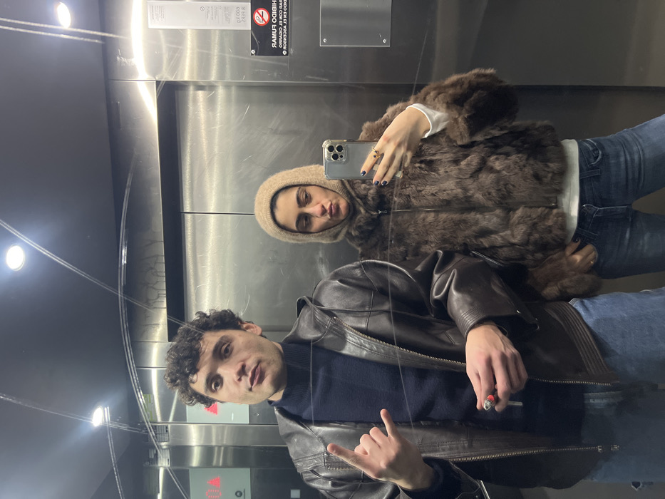 Two people take a selfie in an elevator. The person on the left wears a dark leather jacket over a navy blue sweater and makes a hand gesture. The person on the right wears a fur-like jacket with a hood. They hold up a smartphone and display polished nails. The elevator features a no-smoking sign in Spanish that's partially visible, reflecting their image.