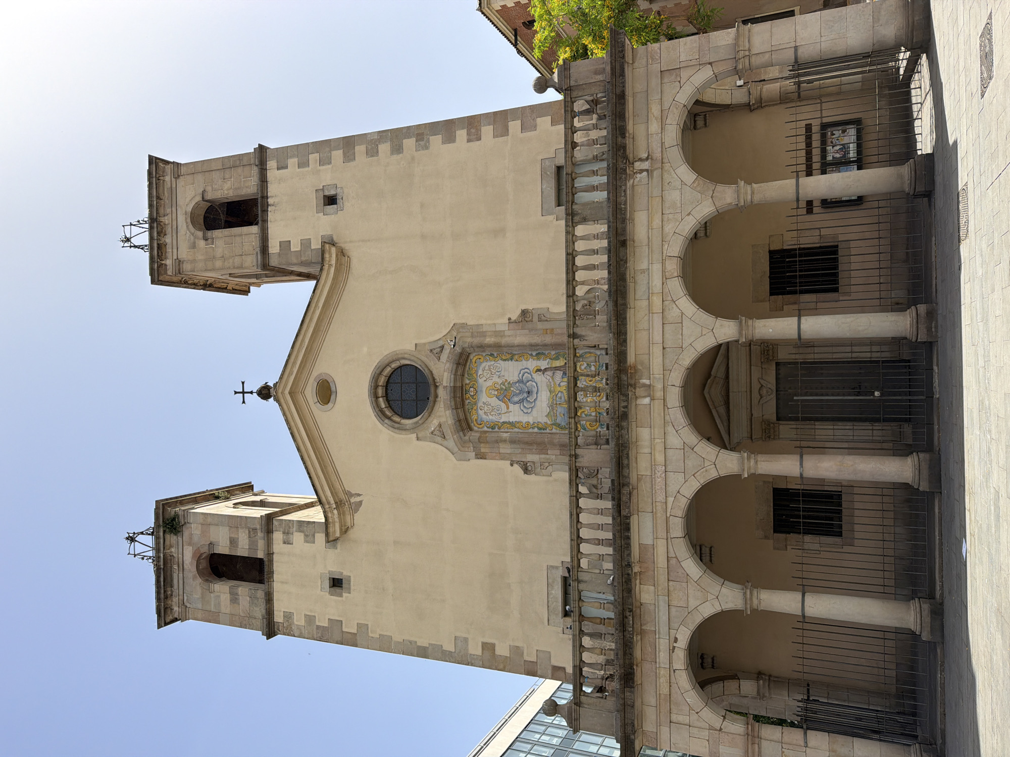 This is a full-frame, eye-level, exterior shot of a church in Barcelona, Spain. The church has a cream-colored facade with two bell towers on either side of the main structure. Both towers are topped with a dark metal structure and a cross. The facade also features a large mosaic depicting a woman holding a baby, surrounded by intricate designs. Below the mosaic is a balcony with a stone railing supported by arched columns. The ground level of the church has a set of large double doors flanked by two arched openings. A black metal fence encloses the entrance area. The sky above is clear and blue. To the left of the church, a modern glass building is visible, and to the right, a building with red brick and a green tree are partially visible. The street in front of the church is paved with stone tiles.