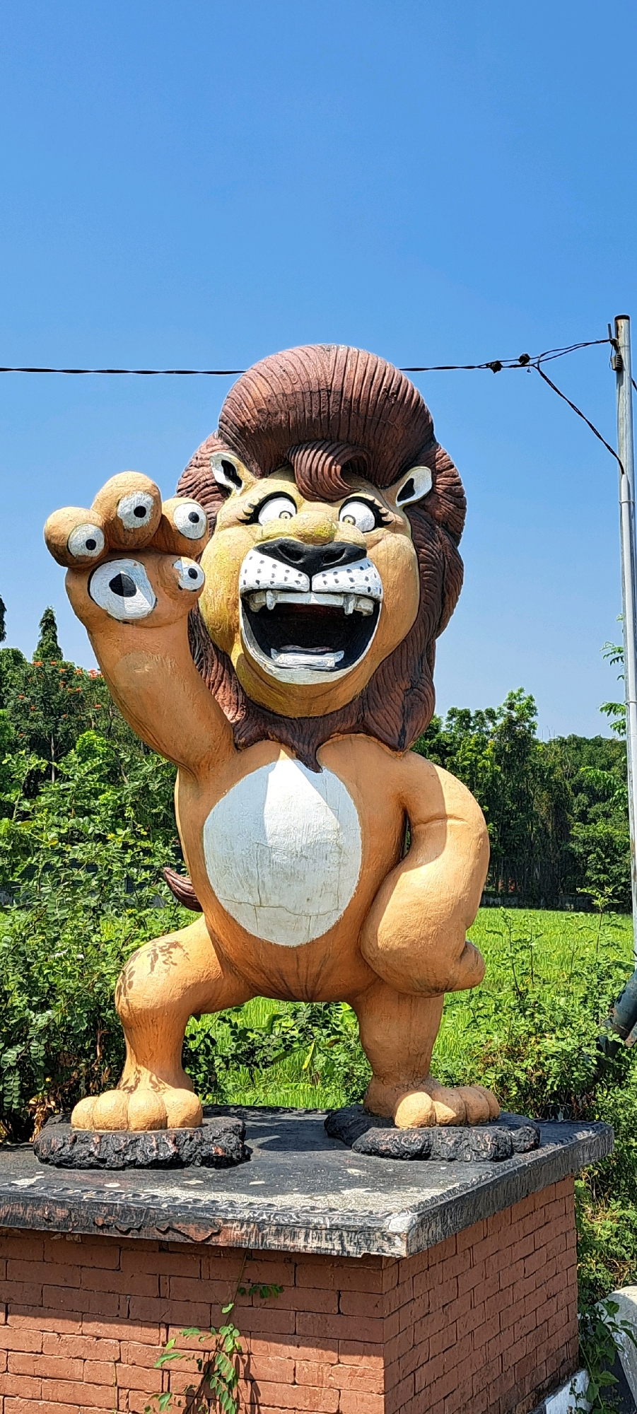 A large, cartoonish lion statue with a brown mane and orange body stands on a brick pedestal. The lion has a wide open mouth with exaggerated teeth and eyes, and its left paw is raised with multiple fingers outstretched. The statue is located outdoors, with a clear blue sky above and lush green foliage surrounding it. A telephone pole and wires are visible in the background, as well as a field of green grass. The location is identified as Lemahbang, Indonesia.