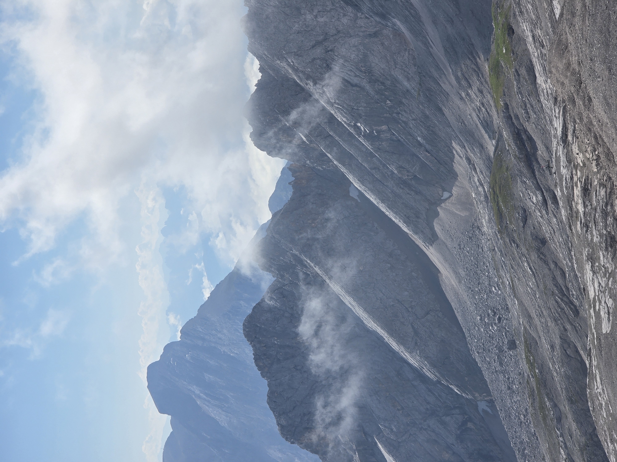 This image captures a mountainous landscape, likely within the Wetterstein Alps near Garmisch-Partenkirchen, Germany. The foreground is dominated by a rocky, scree-covered slope, with patches of sparse vegetation visible on small grassy mounds. Further back, steep, rugged mountain ridges ascend, characterized by sharp peaks and deep ravines. Wisps of white clouds cling to the mountainsides, adding a sense of atmospheric depth and movement. The sky above is a bright blue, with scattered white clouds. In the distance, more layered mountain ranges fade into a hazy blue, suggesting a vast and imposing natural environment. A single figure, appearing as a small silhouette, is visible on the rocky terrain in the mid-ground, indicating the scale of the mountainous terrain.