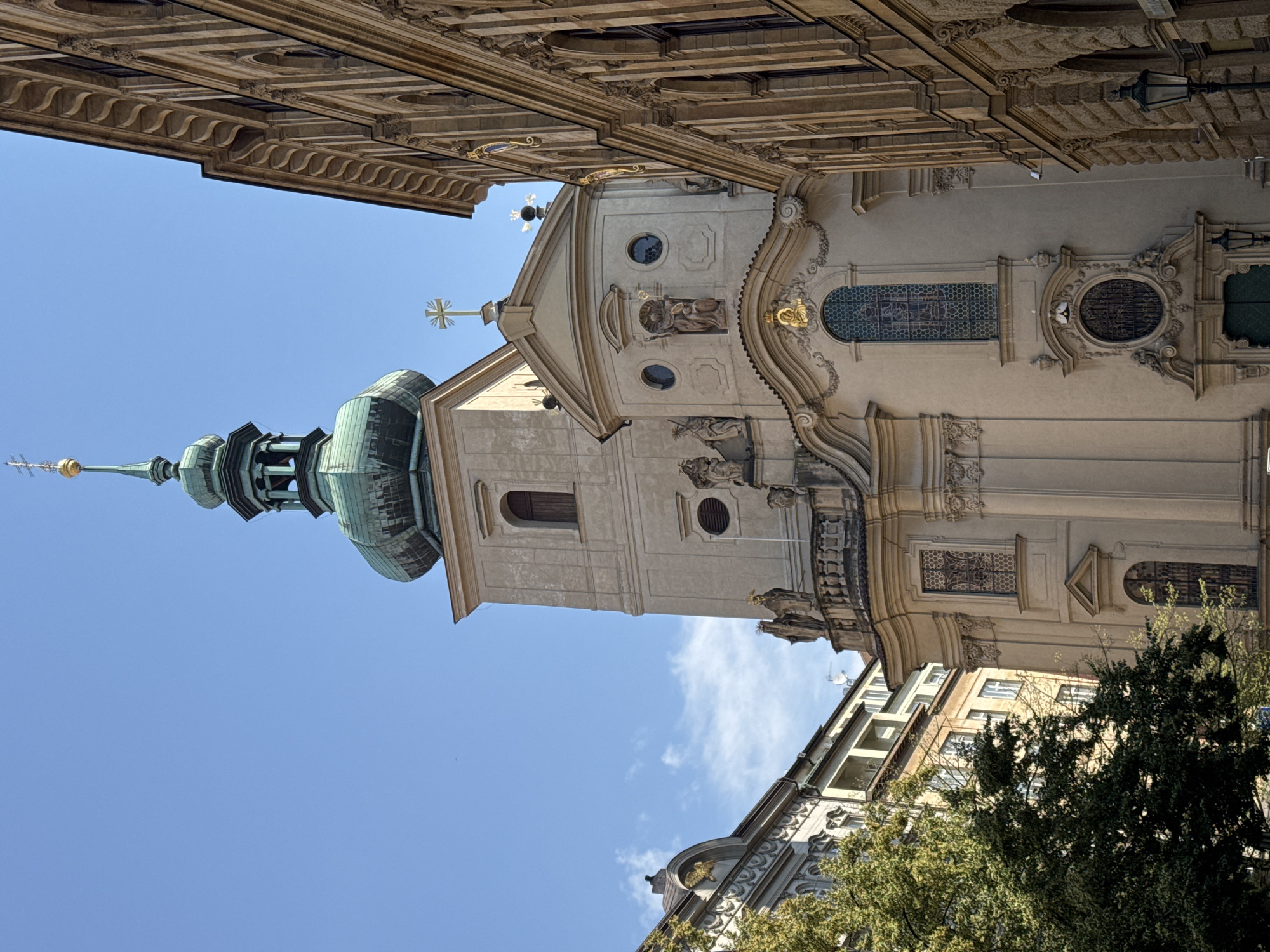 This image captures a detailed view of the Church of St. Nicholas (Kostel svatého Mikuláše) in Prague's Old Town Square, Czechia. The church's Baroque facade is prominent, with its striking white and beige plasterwork, ornate sculptures, and detailed window designs. A tall, slender tower with a green copper dome and a gilded spire rises against a clear blue sky, punctuated by a few wispy clouds. To the right, a section of an adjacent building displays intricate stone carvings and arched windows, typical of Prague's architectural style. The foreground is partially obscured by the lush green foliage of a mature tree, suggesting a street-level perspective. The scene is devoid of people, focusing solely on the magnificent architecture under bright daylight.