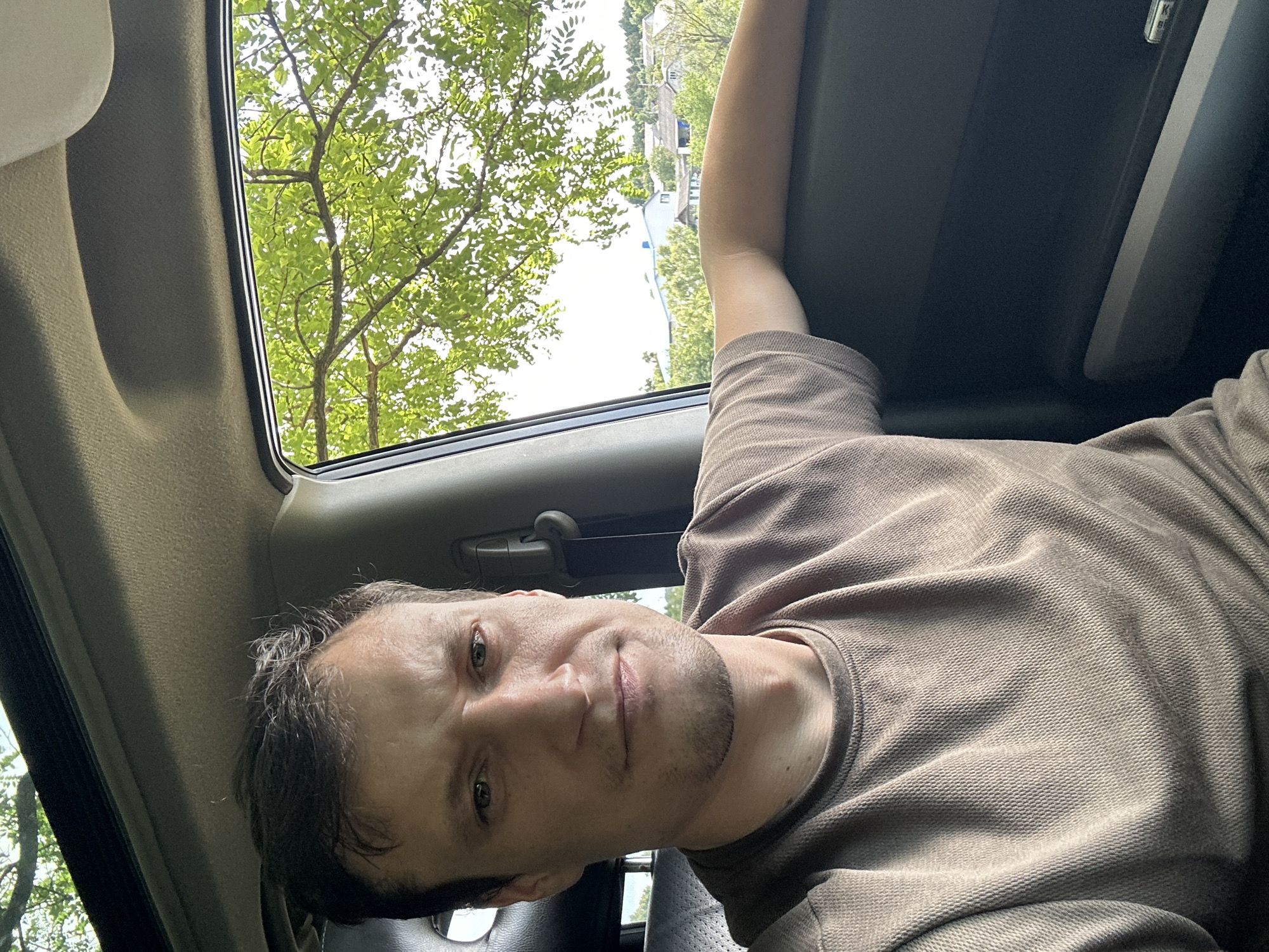 A man is taking a selfie inside a vehicle. He is wearing a brown t-shirt. The man is looking directly at the camera with a serious expression. Through the car window, a tree with green leaves and a few houses can be seen in the background. The scene appears to be in Makiv, Ukraine.