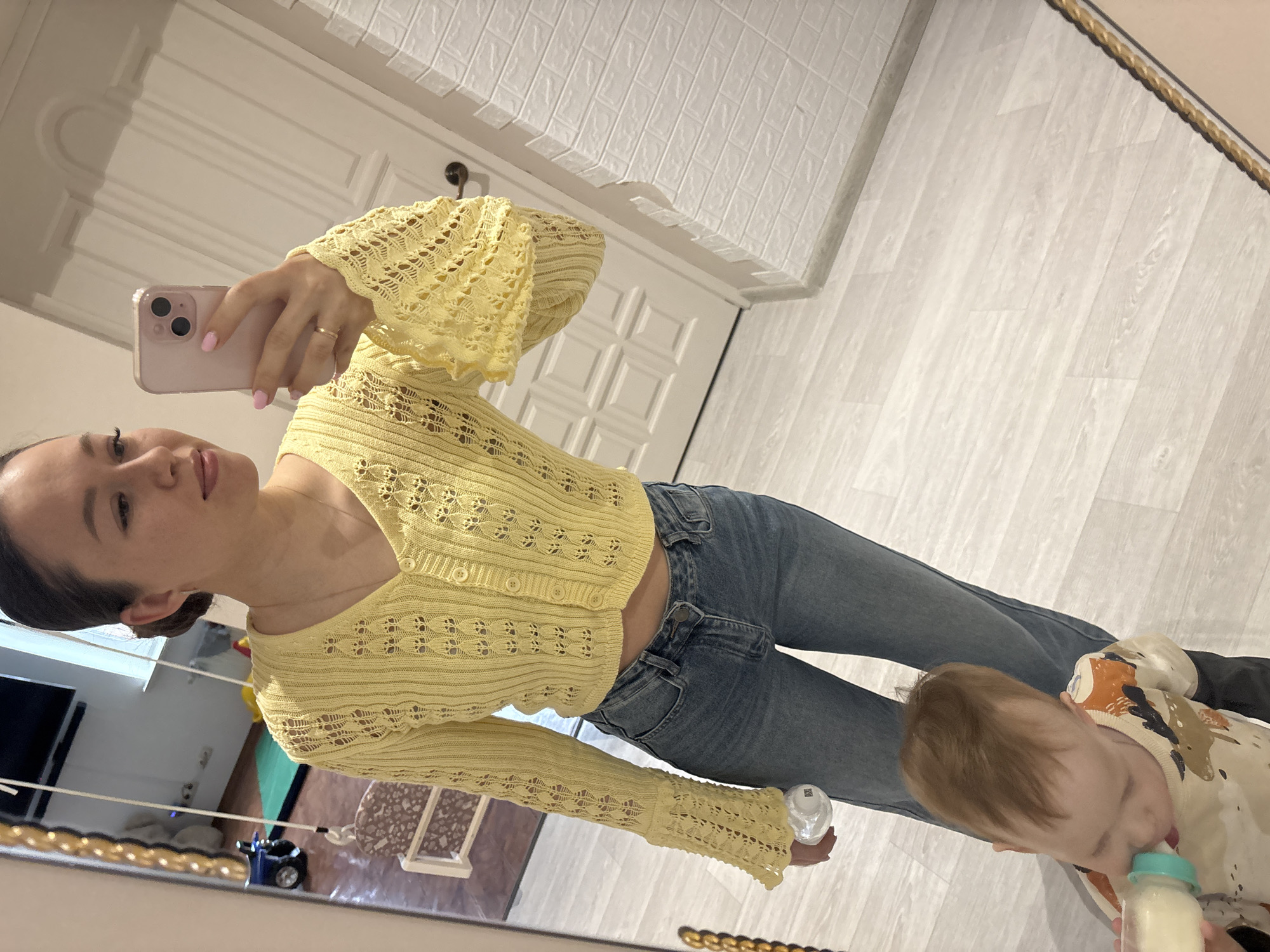 An adult woman is depicted taking a mirror selfie indoors, holding a light pink smartphone with a clear case in her right hand. Her nails are painted pink, and she wears a delicate ring on her right ring finger. She is dressed in a light yellow, long-sleeved, open-knit cardigan with buttons, paired with blue jeans. Her expression is neutral as she looks towards the camera.
Reflected in the mirror, to the lower right, a young child (likely a toddler) is seen from a top-down perspective, focused on drinking from a baby bottle with a light blue/teal nipple. The child wears a light-colored long-sleeved top with abstract patterns in brown, orange, and black.
The background visible in the mirror's reflection includes light-colored wooden laminate flooring and a white paneled door. Above the door, a section of white, textured wall covering, resembling faux brick or tile panels, is visible. Hints of another room or play area with a green mat and a black toy are reflected on the left. The mirror itself is framed by a decorative gold-colored border. The indoor environment appears well-lit. No text is visible in the image. The location context is "Ферма, Russia".