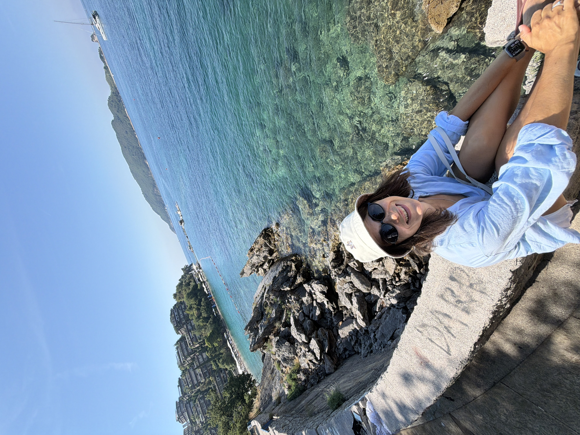 In Boreti, Montenegro, a woman with dark hair, wearing a light-colored bucket hat, sunglasses, and a light blue long-sleeved shirt, sits on a stone ledge overlooking the Adriatic Sea. She is smiling and looking at the camera. The sea is clear and turquoise, with visible rocks and coral beneath the surface. In the background, a line of hotels and apartment buildings is nestled into a green hillside, with a beach and lounge chairs visible in the distance. A white sailboat is anchored in the bay. The sun is shining brightly, casting shadows on the stone ledge where the woman is seated. The water is calm and inviting. The overall scene depicts a sunny day at a coastal resort.