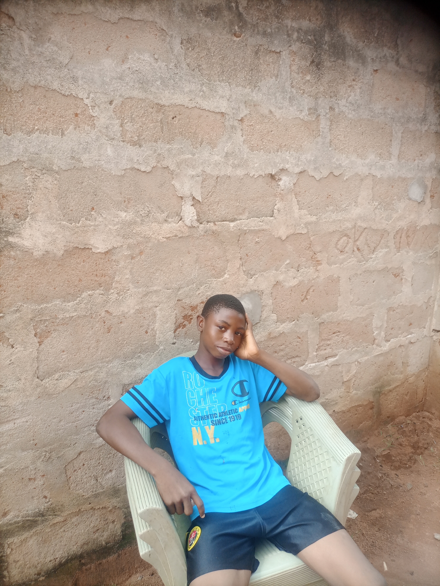 In Nkpor, Nigeria, a young boy with dark skin and short black hair sits in a light-colored plastic chair against a rough, textured concrete wall. He is wearing a bright blue t-shirt with white text and graphics, including the word "ROCHE" and "N.Y.", and black shorts. His left arm is bent, with his hand resting on his head, and his right arm is also bent, resting on the armrest of the chair. The wall behind him is a dull gray and tan color, with visible blocks and rough plastering. The ground beneath the chair appears to be dirt with some sparse vegetation. The overall scene is outdoors, under natural lighting.