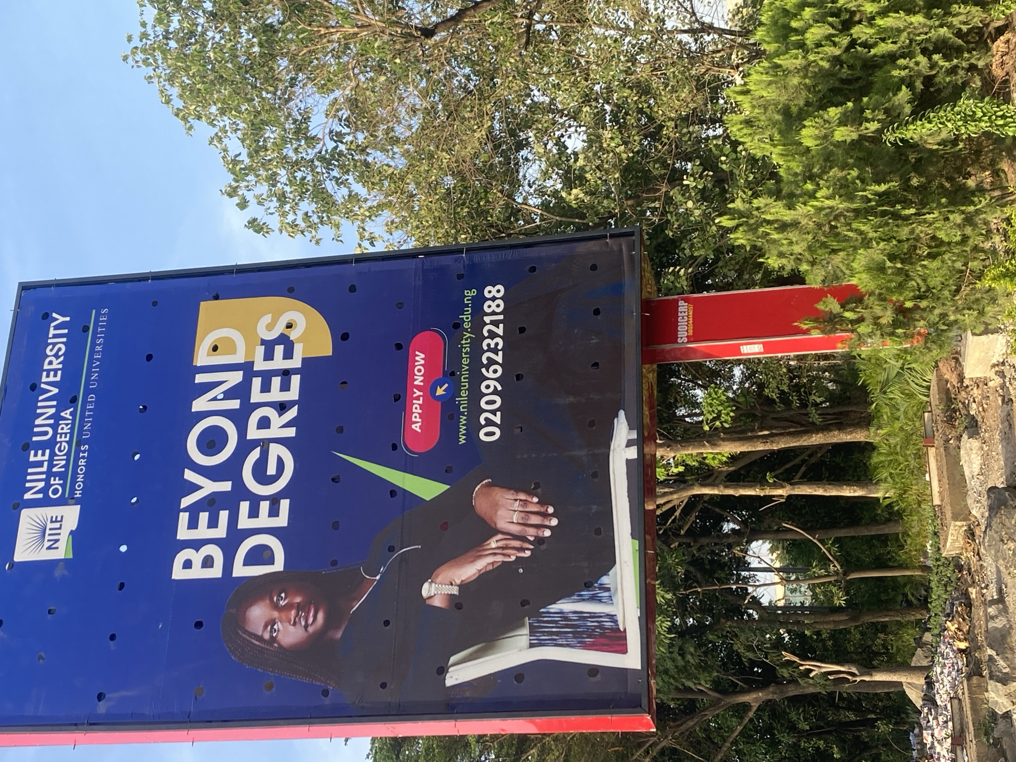 The image shows a roadside billboard for Nile University of Nigeria. The billboard has a blue background with white and yellow text, as well as an image of a woman. The text reads "NILE UNIVERSITY OF NIGERIA, HONORIS UNITED UNIVERSITIES, BEYOND DEGREES, APPLY NOW, www.nileuniversity.edu.ng, 02096232188". A part of a red sign that read "SUOICERP 08054444057" is also visible. The billboard is surrounded by trees and some ground debris. Based on the information on the billboard, the scene is located somewhere in Nigeria. It is unclear from the image whether the location is actually in Karmajiji, Nigeria.