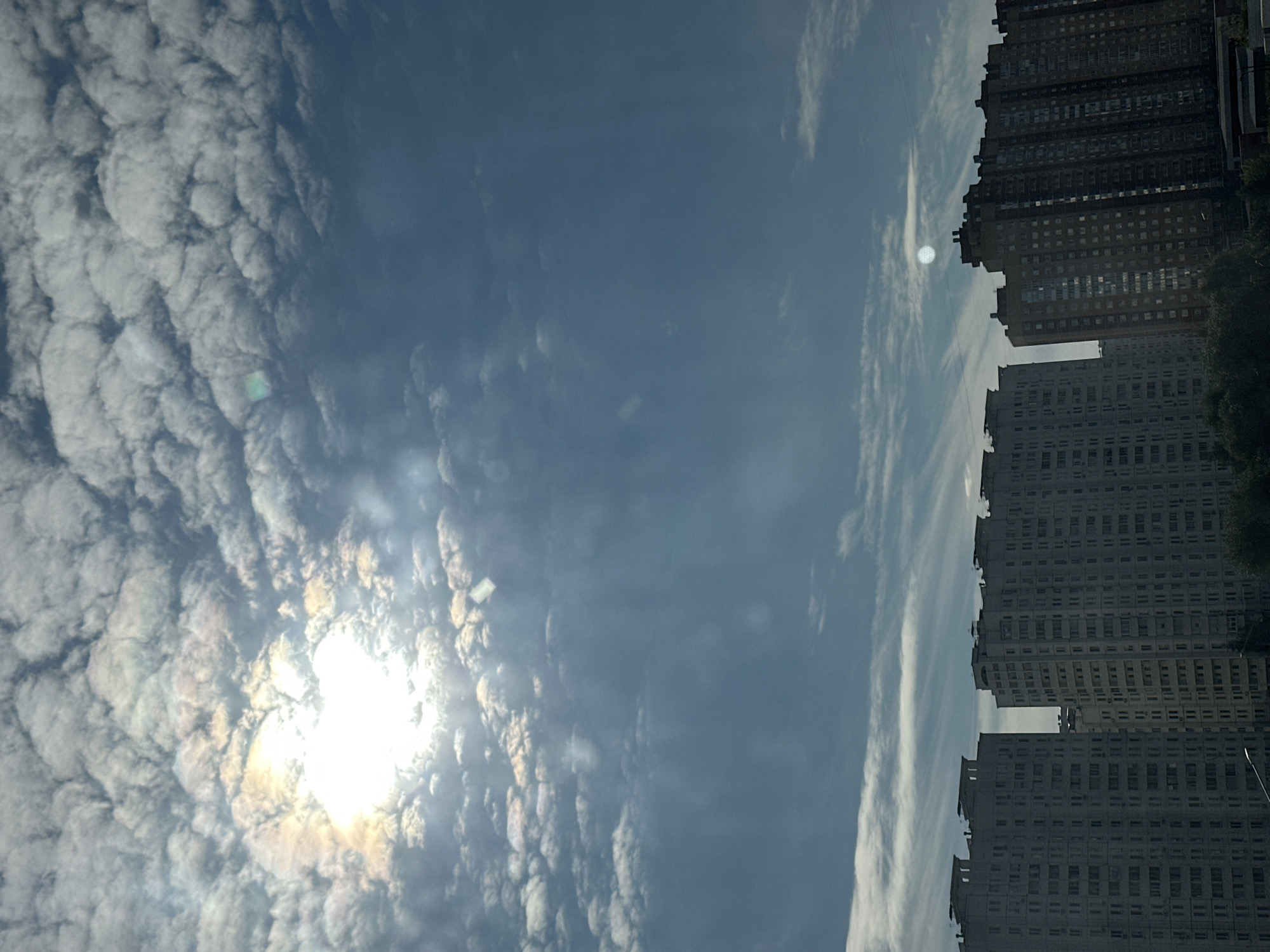 A view of apartment buildings under a cloudy sky with the sun visible. The buildings are tall and appear to be residential. Some are light-colored and monolithic, while others are darker with a brick-like facade. Wispy clouds are scattered across a blue sky, with the sun creating a bright glare. The image is taken from a distance, looking up at the buildings.