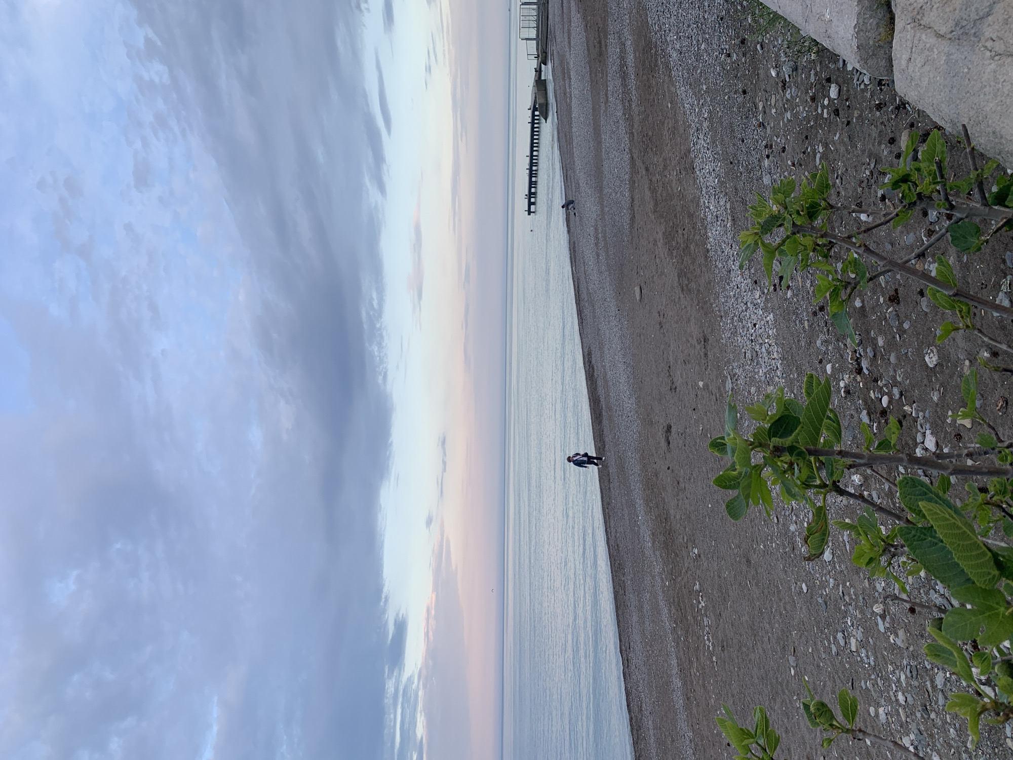 The image shows a coastal scene with a beach, ocean, and sky. The beach is to the right and appears to be composed of sand and small pebbles. Some green foliage is visible on the right edge of the frame. The ocean stretches out to the left, leading to a distant horizon line. In the distance, a structure that resembles a pier or jetty is present. One person is walking on the edge between the beach and the water. The sky is covered with clouds, showing a range of blue and grey shades, possibly indicating early morning or late evening light. The image appears to be taken at a slightly canted angle.