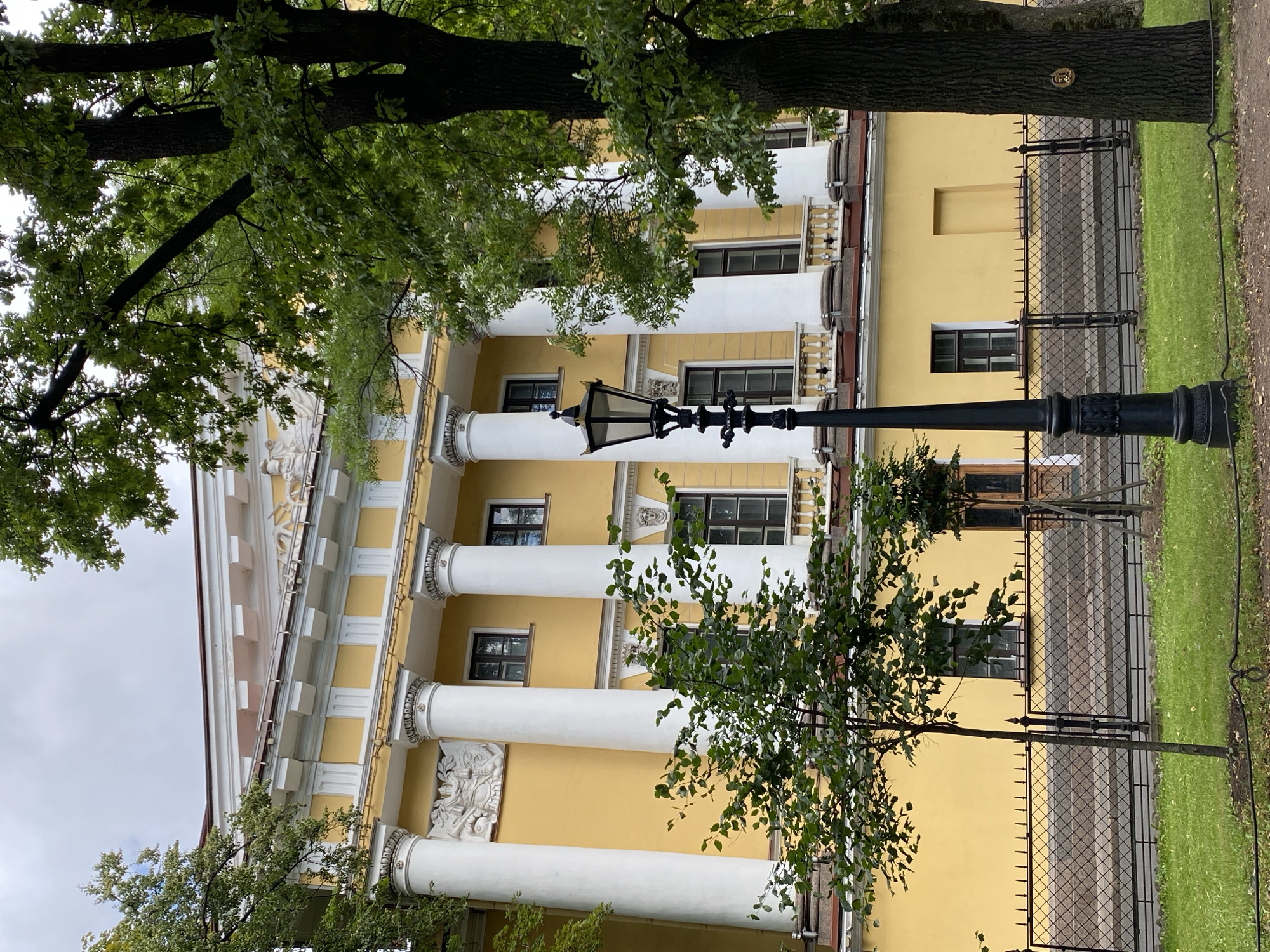 This is a photograph of the Saint Petersburg State University of Economics (UNECON) building, located at 1/5, Sadovaya Street, Saint Petersburg, Russia. The image shows the yellow facade of the neoclassical building, characterized by large white Ionic columns and ornate decorative elements. In the foreground, a black lamppost stands prominently, and a low black fence separates the building from a grassy area. Mature trees with lush green foliage partially obscure the view of the building and the sky, which is overcast. The composition is framed by the branches and leaves of these trees, suggesting an outdoor setting on a cloudy day. There are no people visible in the image.