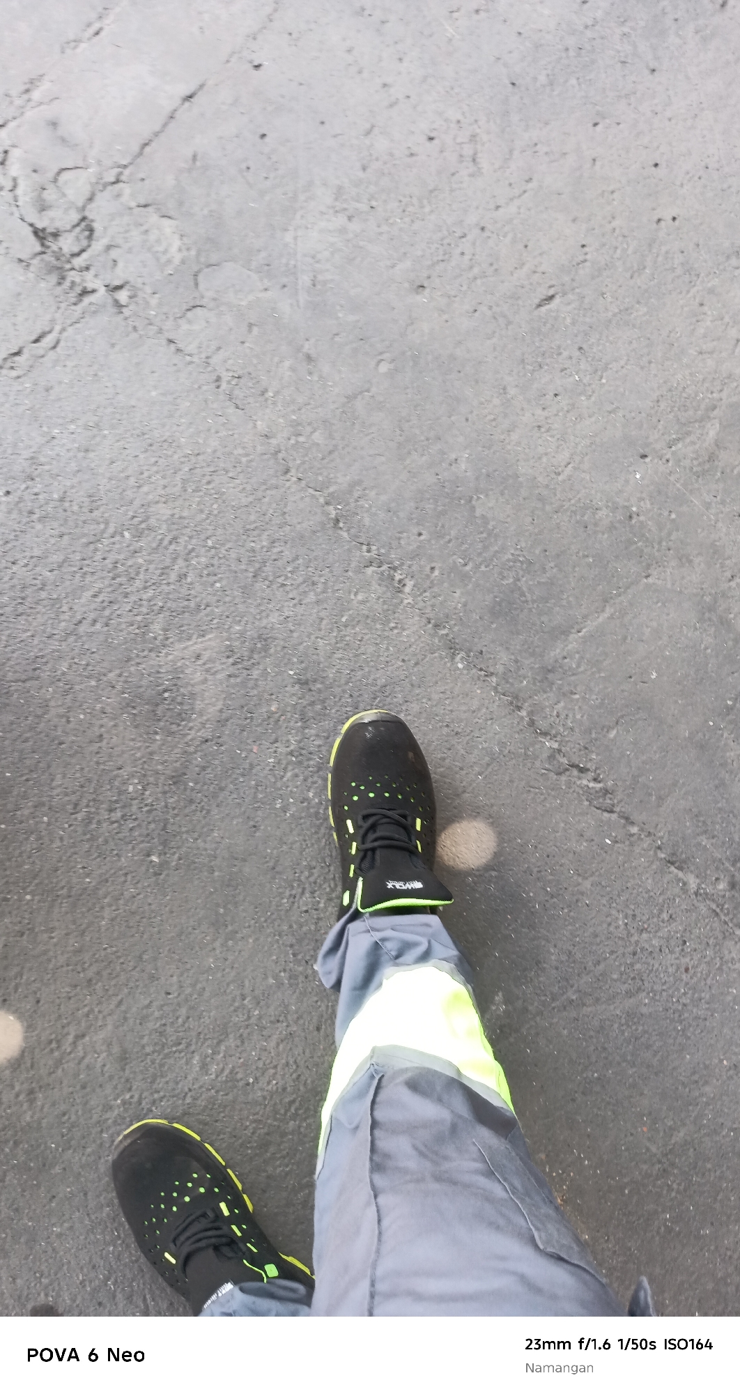 The image shows a first-person perspective of someone standing on a cracked asphalt surface. The subject is wearing dark athletic shoes with neon green accents and gray work pants with neon yellow reflective stripes. The camera information at the bottom indicates the photo was taken with a POVA 6 Neo phone, at 23mm focal length, f/1.6 aperture, 1/50s shutter speed, and ISO 164. The location is identified as Namangan City, Uzbekistan.