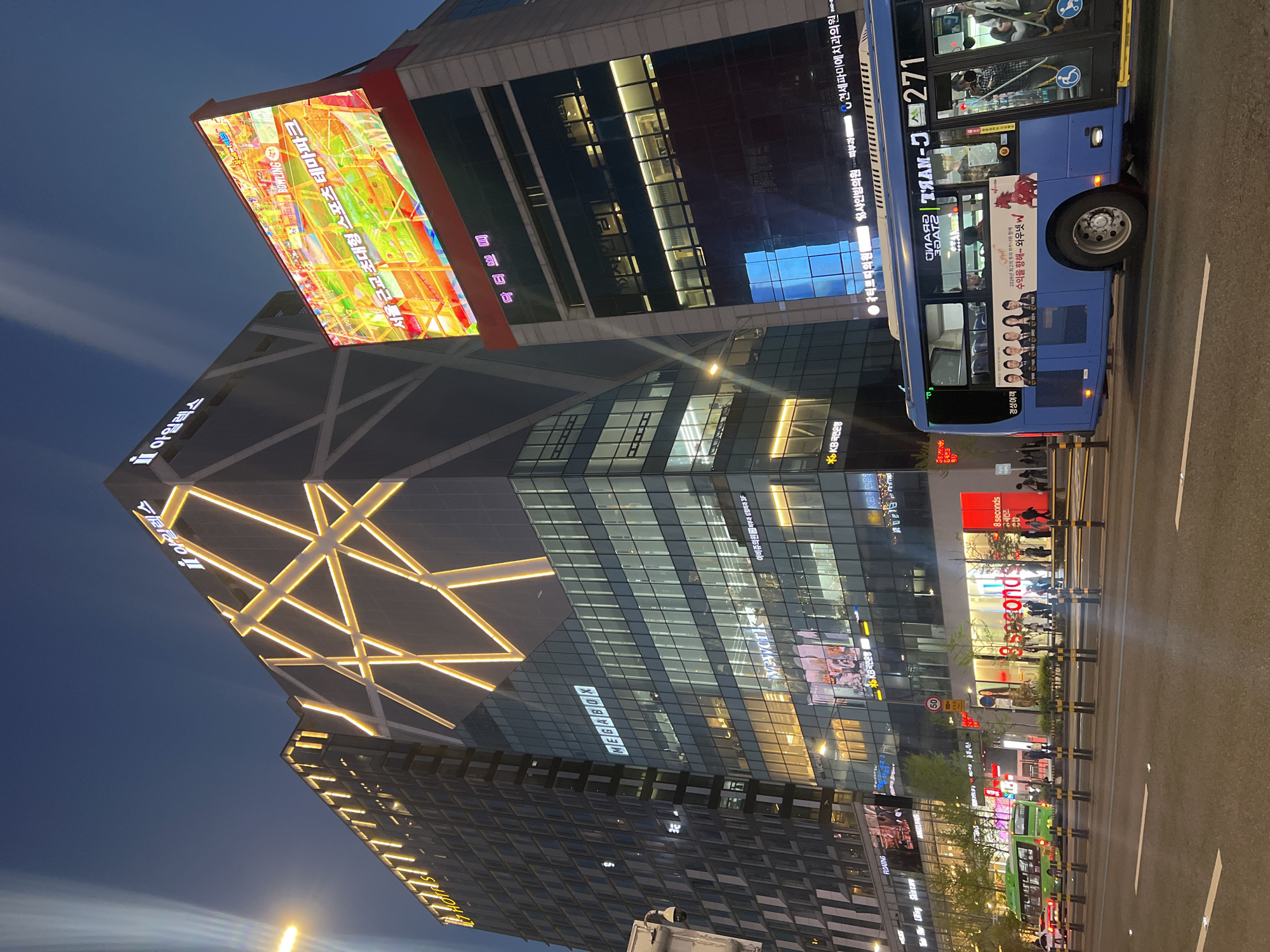 This is a night scene in Seoul, South Korea. A tall, modern building dominates the view. Its exterior features a triangular geometric design with illuminated golden lines. Atop the building is a large LED screen displaying colorful content. The building’s façade is also adorned with signs, including "아일렉스" and "MEGABOX." Street-level storefronts, including "8 seconds," are visible at the base of the building. A blue bus labeled "7271" is parked on the street. Multiple pedestrians are visible in the background. The time appears to be twilight or night, indicated by the dark sky and lit signage.