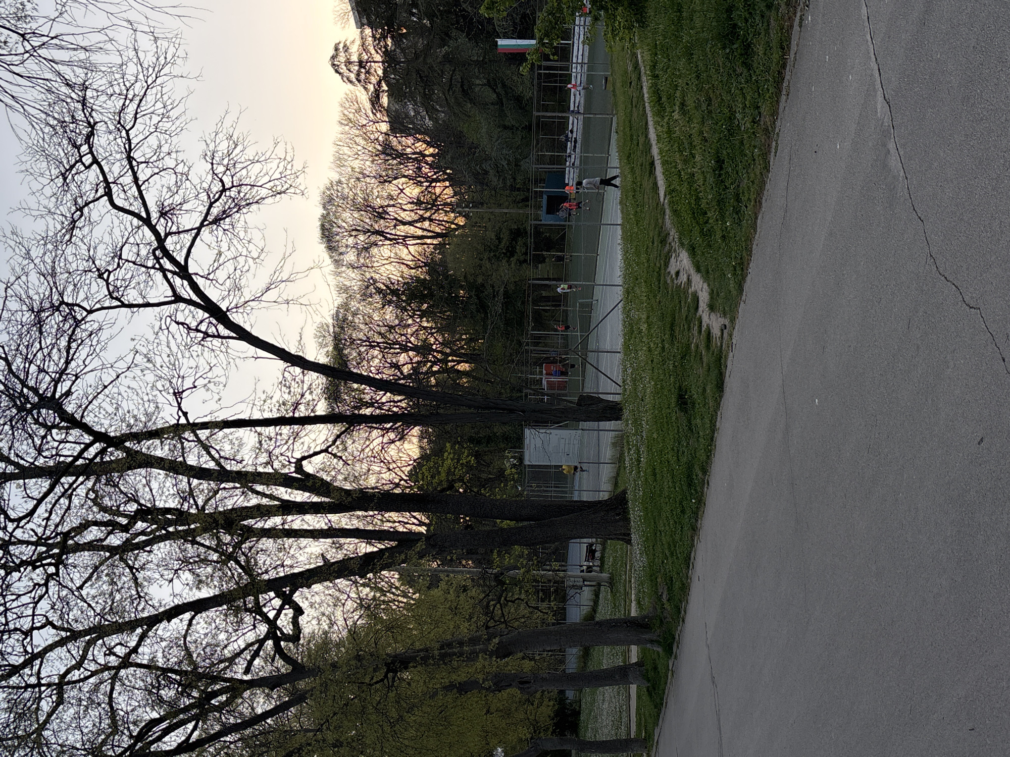 A sideways, low-angle shot taken in the Sea Garden in Varna, Bulgaria, captures a paved pathway lined with trees, a chain-link fenced area, and individuals engaged in activities.
The trees are diverse, with some still bare while others show early signs of leaves. A gray, cracked paved path runs along the right side. Adjacent to the path is grass interspersed with small white flowers. Behind the trees on the right, a chain-link fence encloses what appears to be a sports area, possibly a tennis or basketball court, where several people are engaged in various activities.
In the background, the sky is muted, suggesting twilight or early evening. A flag is visible, indicating the location is likely Bulgaria.