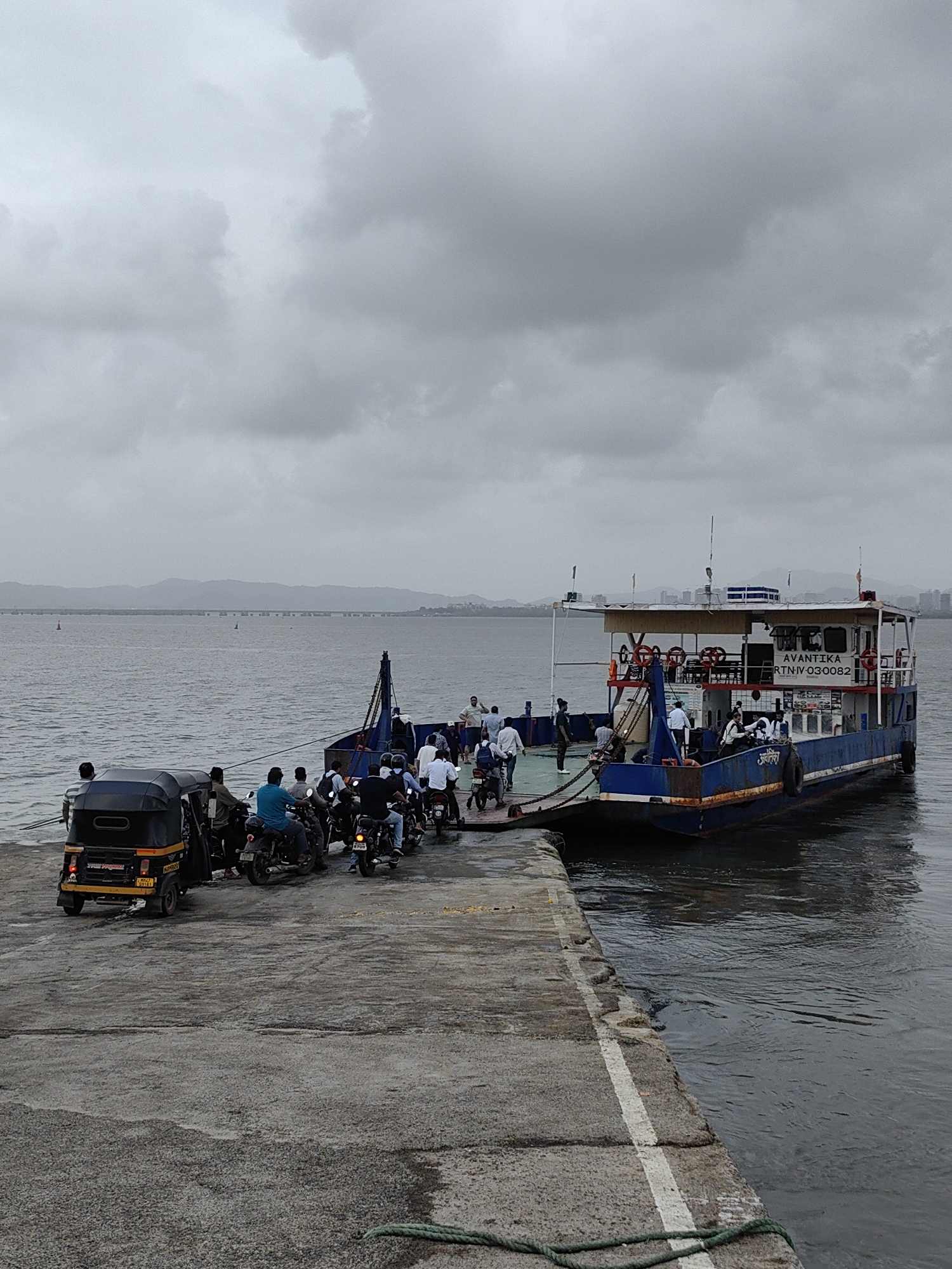 A ferry boat, named "AVANTIKA," is docked at a concrete pier on a cloudy day. The ferry is blue and white with "AVANTIKA" and "RTN.IV-030082" printed on its side. Several people are visible on the ferry and on the pier. On the pier, an auto-rickshaw and several motorcycles are lined up, suggesting a ferry service for vehicles. The water is calm, and a distant cityscape with buildings and a bridge is visible across the bay. The scene appears to be in Mira-Bhayander, India, a coastal city known for its ferry services across the Thane Creek.