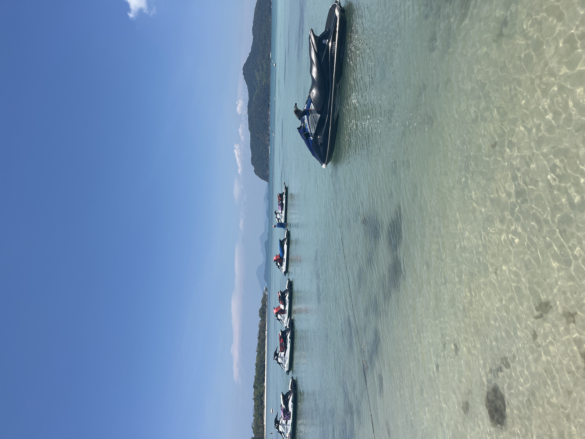 The image depicts a coastal scene near Baan Thong Krut, Thailand, on a sunny day. Several jet skis are lined up in the clear, shallow water near the shore. One black jet ski, occupied by a person, is positioned further out from the others. In the background, land is visible, featuring hilly terrain covered with vegetation. The sky is mostly blue with a few scattered clouds. The water is clear enough to see the sandy bottom.