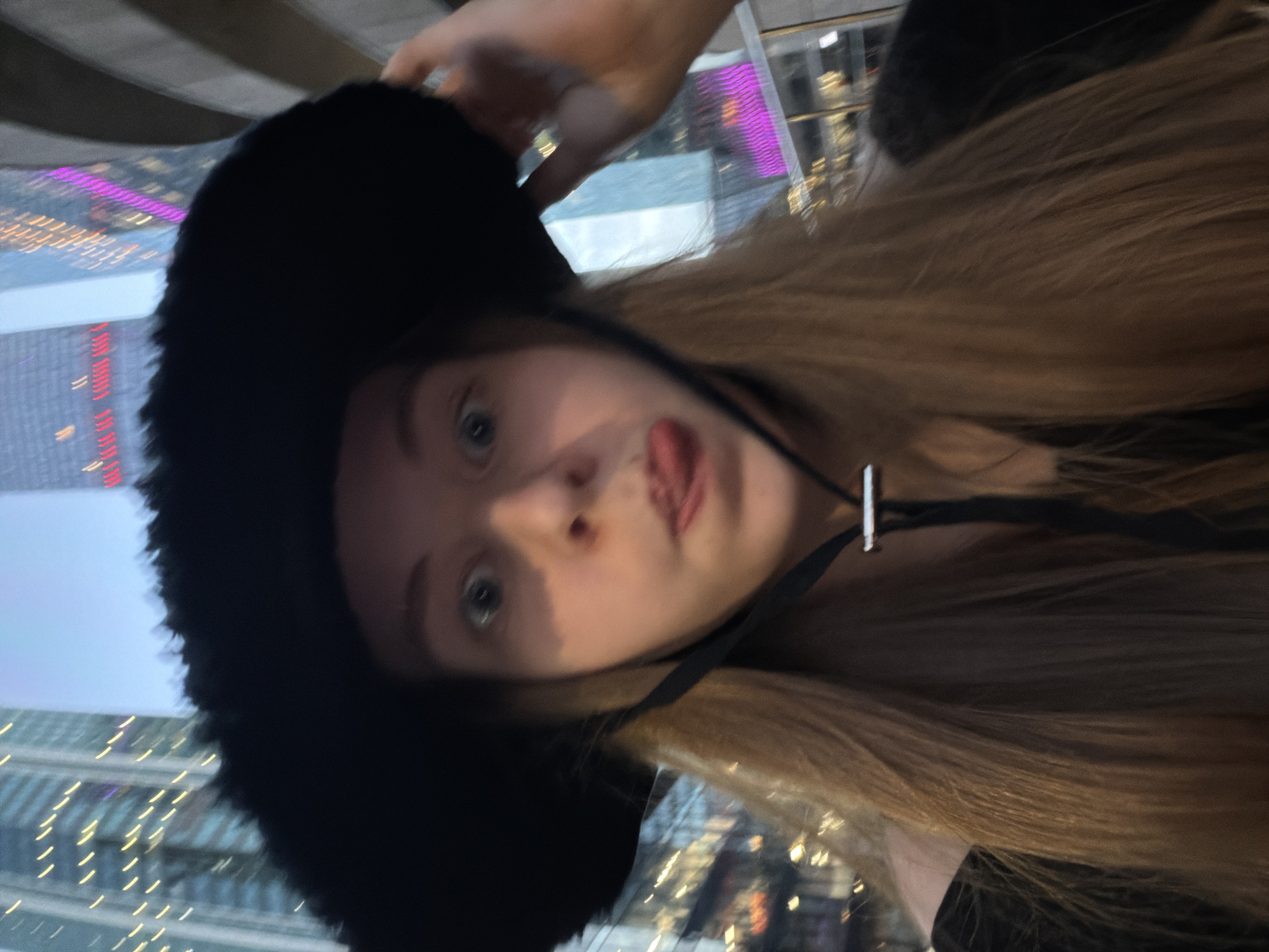A young woman with long, blonde hair and light-colored eyes is shown from the chest up. She is wearing a black, fuzzy, wide-brimmed hat with a black string tie. Her mouth is open, and her tongue is sticking out. The background appears to be a city at dusk or night, with tall buildings illuminated by lights. The buildings in the background suggest an urban environment, possibly a commercial or business district. The specific location of Khimki, Russia is not explicitly indicated by any visual cues in the image.