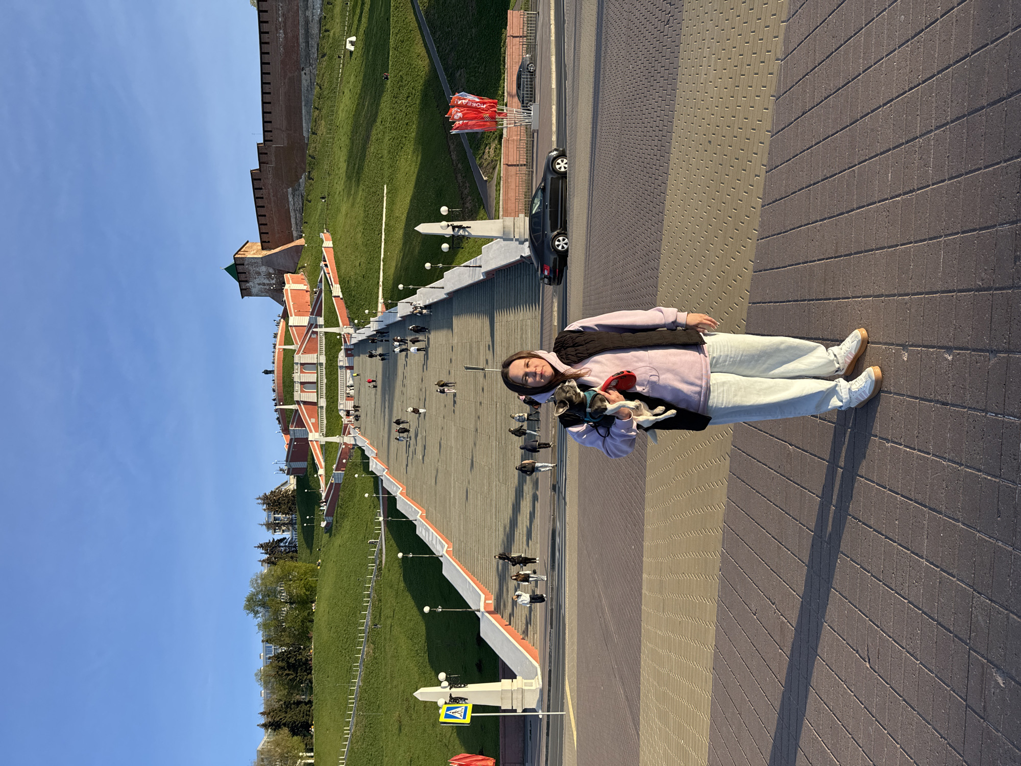 A young woman stands holding a dog in front of the Chkalov Stairs in Nizhny Novgorod, Russia, on a sunny day. The stairs lead to a building or structure at the top of the hill. People are walking up and down the stairs. A car is parked nearby. The stairs are bordered by green grass and lamp posts. A crosswalk sign is visible in the foreground. The sky is blue.