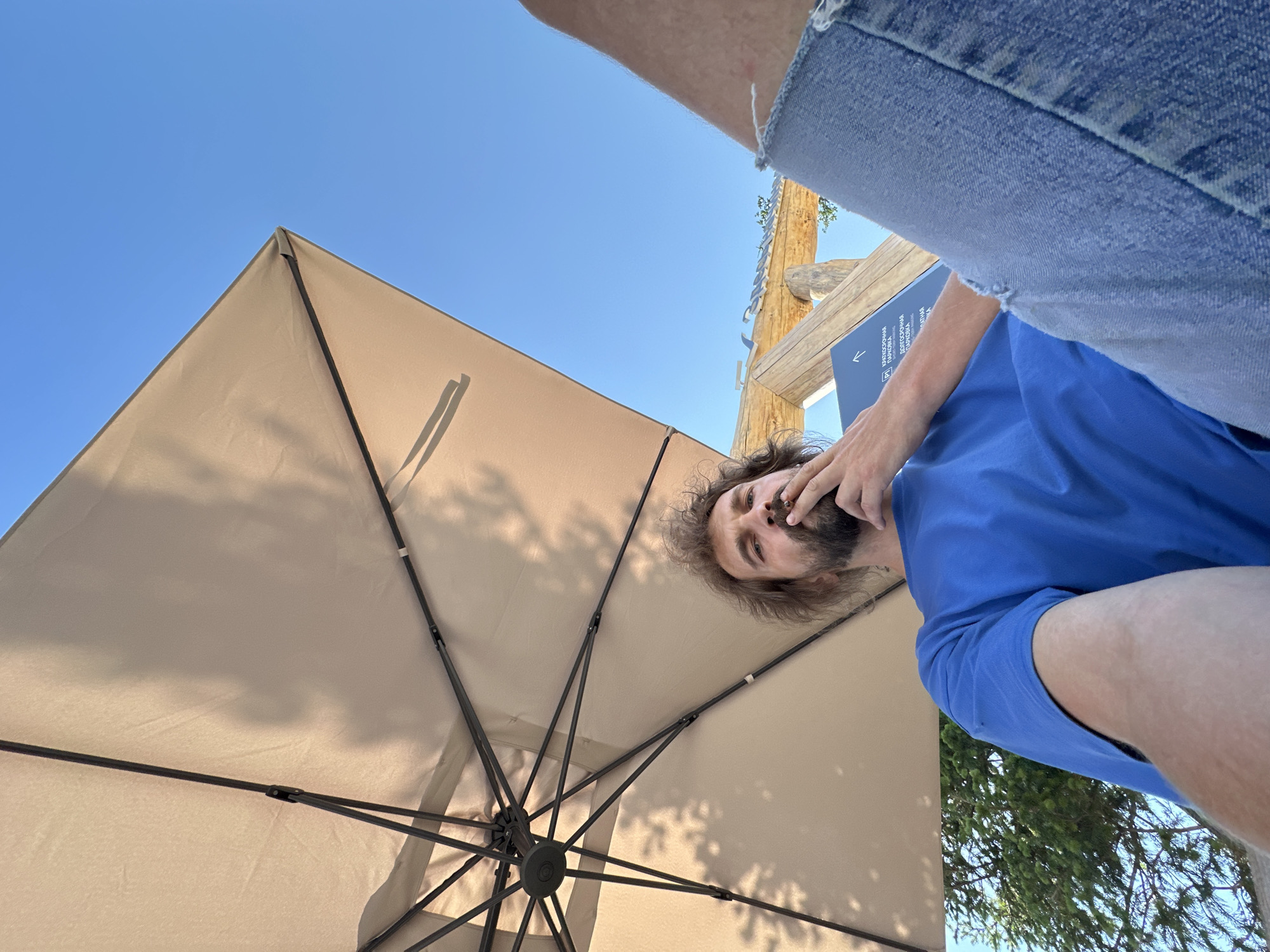 A low-angle, wide shot captures a man sitting outdoors under a large beige umbrella. The man, with long, unkempt brown hair and a beard, is wearing a blue t-shirt and ripped denim shorts. He is holding a cigarette to his lips and appears to be smoking. The umbrella's frame and fabric are visible, with shadows from trees cast on its surface. In the background, a clear blue sky is visible, along with a wooden structure and some greenery. A blue sign with white text is partially visible on the wooden structure. The image seems to be taken in Maima, Russia, given the context provided.