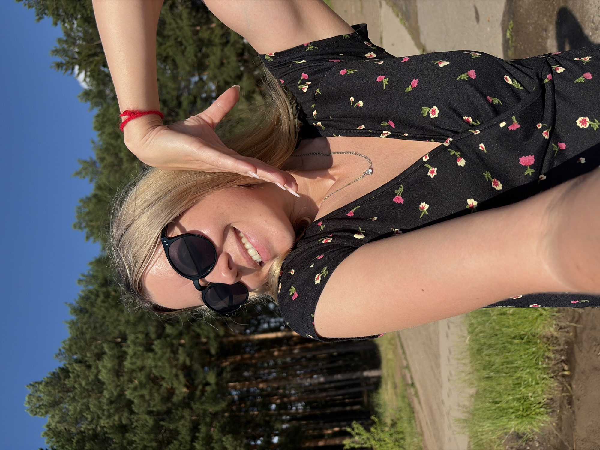 A woman with blonde hair and sunglasses is taking a selfie outdoors. She is wearing a black dress with a floral pattern and a silver necklace. She is smiling and making a heart shape with her hands. The background shows a forest and a clear blue sky. The image appears to be taken in Chaikovsky, Russia, given the presence of tall evergreen trees and a dirt path, consistent with the landscape of the region.