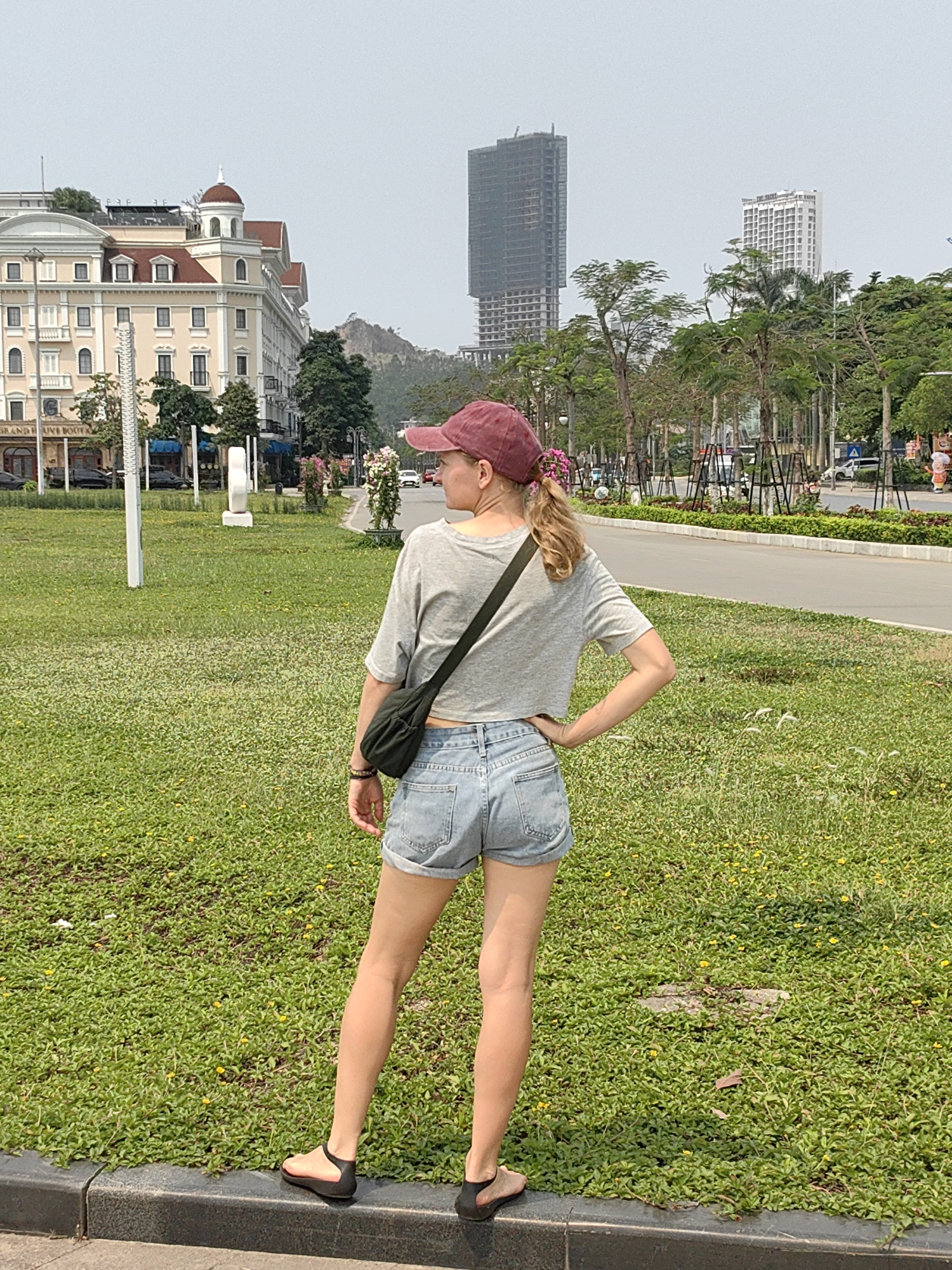 A woman stands on a curb, facing away from the viewer, in a city park in Bai Chay Ward, Vietnam. She wears a maroon baseball cap, gray cropped t-shirt, jean shorts, and black sandals, with a black bag slung over her shoulder. Her left hand rests on her hip.
Behind her, a large green lawn dotted with small yellow flowers stretches towards a sidewalk and a road. Across the road is a row of trees and shrubs. In the background, several buildings are visible, including a large, ornate hotel on the left, and two tall, modern skyscrapers further back. A small mountain is visible behind the buildings.