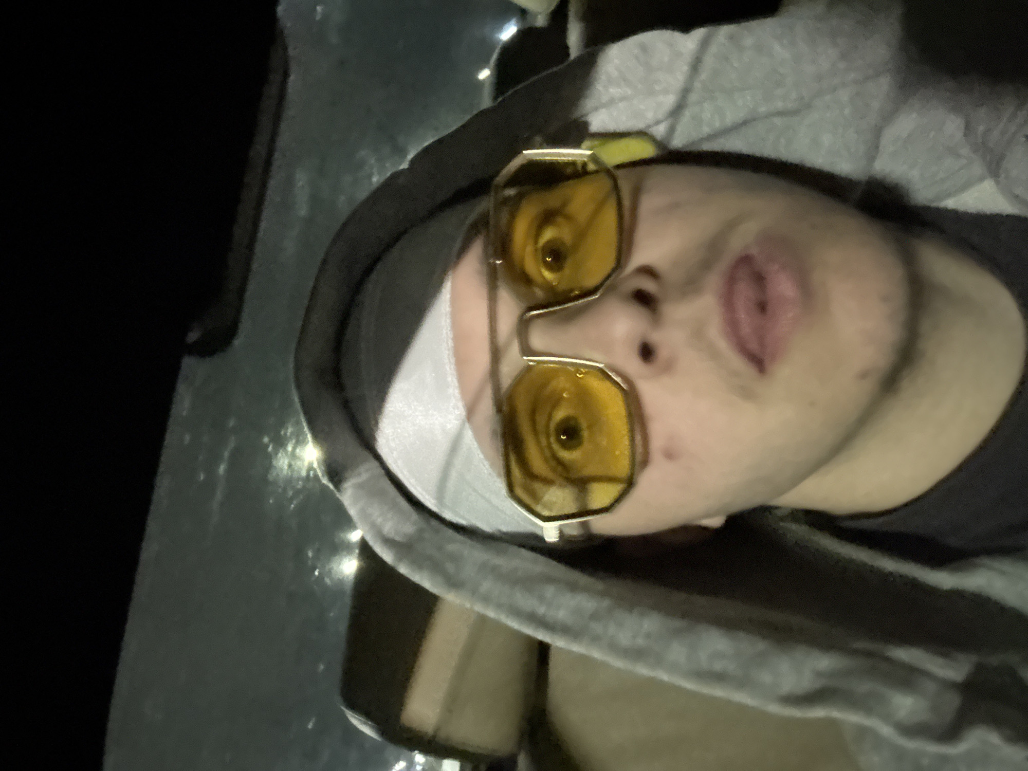 A person is seen in a close-up, low-angle shot. The individual is wearing a gray hooded sweatshirt and a white headband. They have on a pair of yellow, octagonal-shaped sunglasses. The subject's facial expression suggests they may be sticking their tongue out slightly. The background is dark, with a reflective surface behind the person, possibly a car window. There is no indication of a specific location in Sofia, Bulgaria.