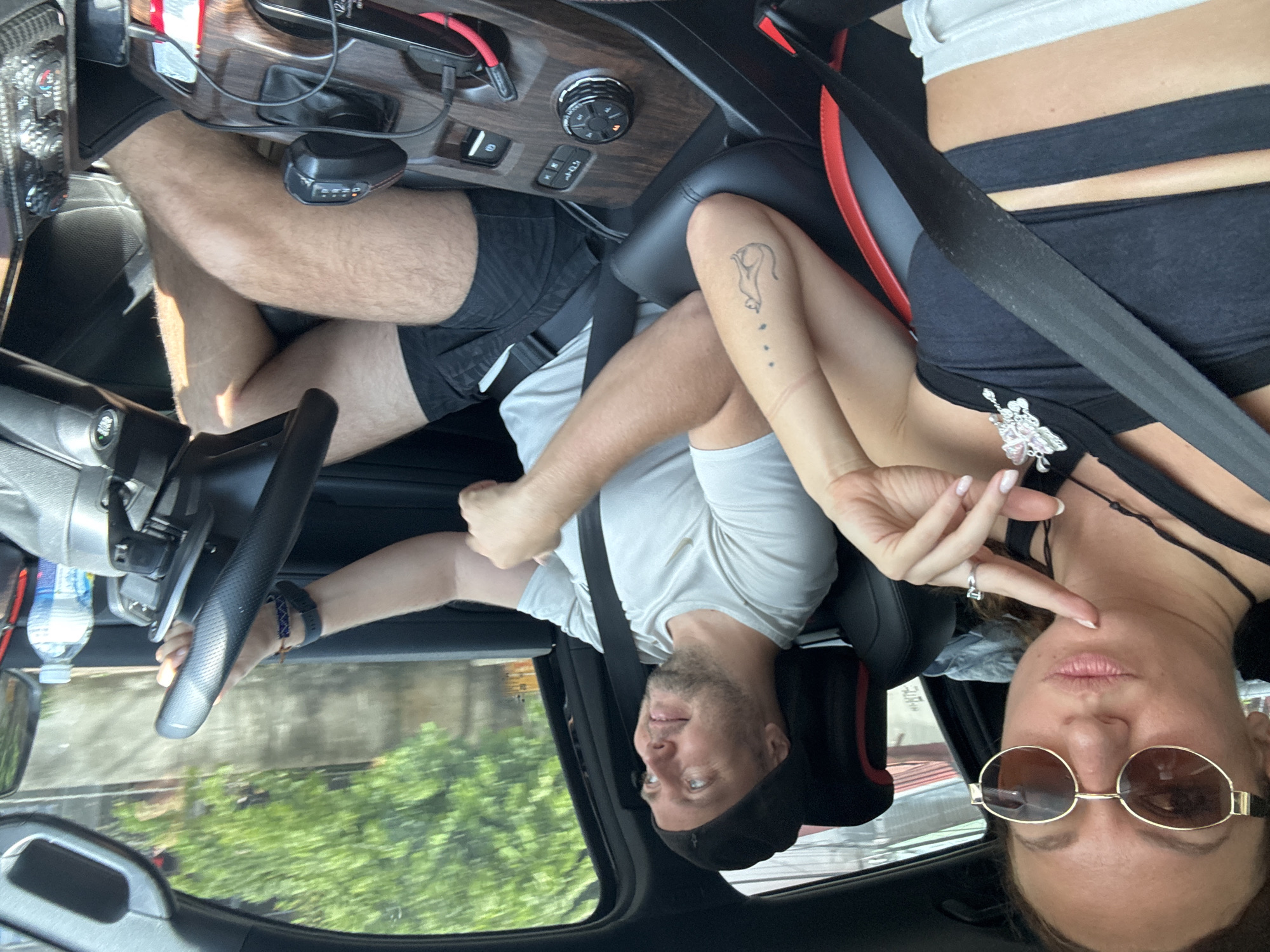 Inside a vehicle, a man and a woman are visible. The man is in the driver's seat, wearing a white t-shirt and a black cap. The woman is in the passenger seat, wearing a black top and sunglasses. They are both wearing seatbelts. The car's interior is visible, including the dashboard and steering wheel. Outside the car, trees and foliage suggest a natural environment. Based on the instructions, the location of the image is Mèo Vạc Commune, Vietnam.
