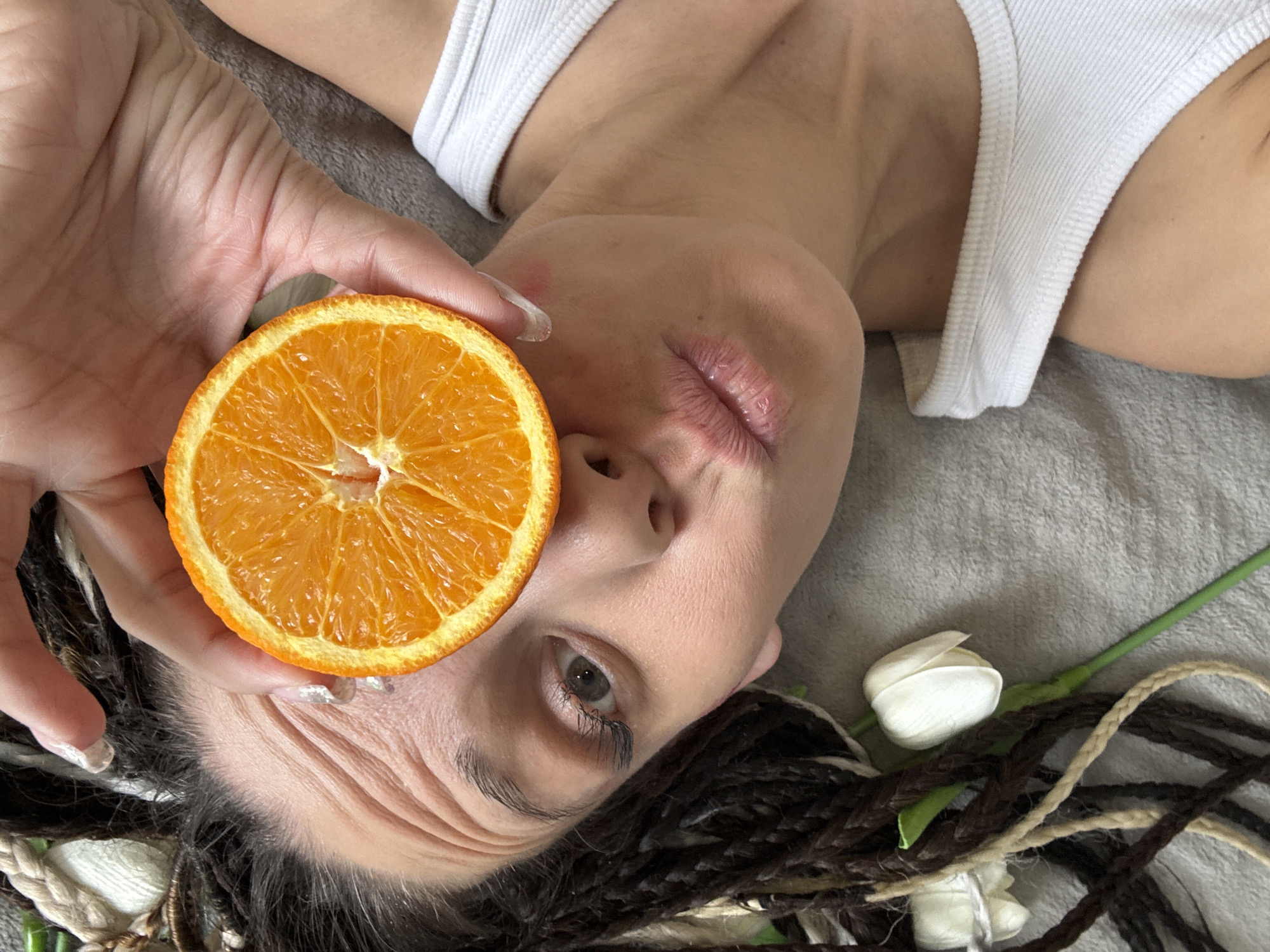 A woman with braided hair, wearing a white tank top, is lying on a gray surface. She holds a half-cut orange in her right hand near her face. A white flower lies near her hair on the gray surface. The location is Unknown, Unknown.