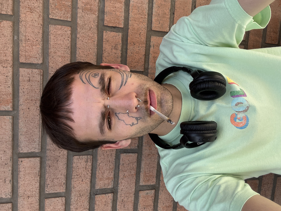 A person is depicted from an overhead perspective, lying down with their head turned to the right. The individual appears to be male, with short, dark brown hair styled with bangs. Their face is adorned with several piercings, including a barbell piercing above one eyebrow and multiple rings in their nose. Distinctive tattoos are visible on their face: intricate swirl patterns near their temples and a map-like outline on their right cheek/jawline. They have stubble on their chin and cheeks. A cigarette or joint with a lit ember is held between their lips.
Around their neck are black over-ear headphones. They are wearing a light green t-shirt. On the chest of the t-shirt, a colorful graphic is partially visible, showing the letters "GOL" in various vibrant hues (orange, blue, green, pink, yellow). The background consists of a reddish-brown brick wall, suggesting the person is lying either on the ground or a surface adjacent to the wall. The lighting is bright and natural, indicating it is daytime. The person's eyes are partially closed, contributing to a relaxed or contemplative expression.