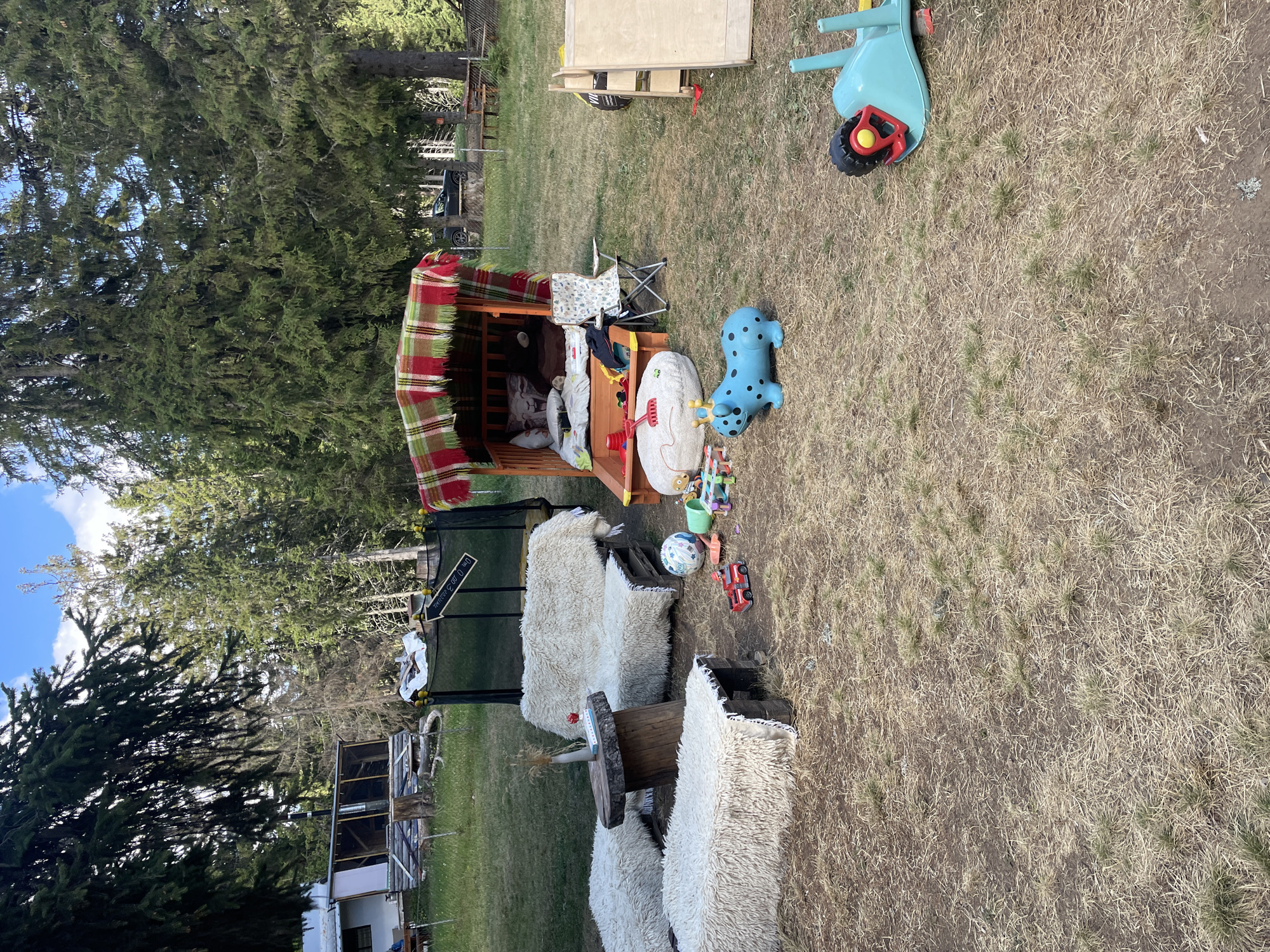 The image depicts an outdoor seating area and play space in a rural or backyard setting, possibly near Smolyan, Bulgaria. In the foreground, there are two low, rectangular benches and a larger L-shaped sofa, all covered with shaggy, off-white material. A rustic wooden table sits between the L-shaped sofa and one of the benches. Scattered on the ground are various children's toys, including a red toy truck, a soccer ball, a blue toy hippo with spots and a crown, and a green toy rake.
Further back, a covered wooden structure, possibly a playhouse or fort, is set up with bedding inside and a colorful plaid blanket draped over it. A small folding chair is placed next to this structure. A trampoline is visible in the background, partially obscured by trees. To the right, a blue toy wheelbarrow lies on its side. The ground is covered with dry grass and dirt. In the far background, trees dominate the landscape, and hints of buildings and a car suggest a residential or recreational area. The overall impression is one of a whimsical, DIY play area.