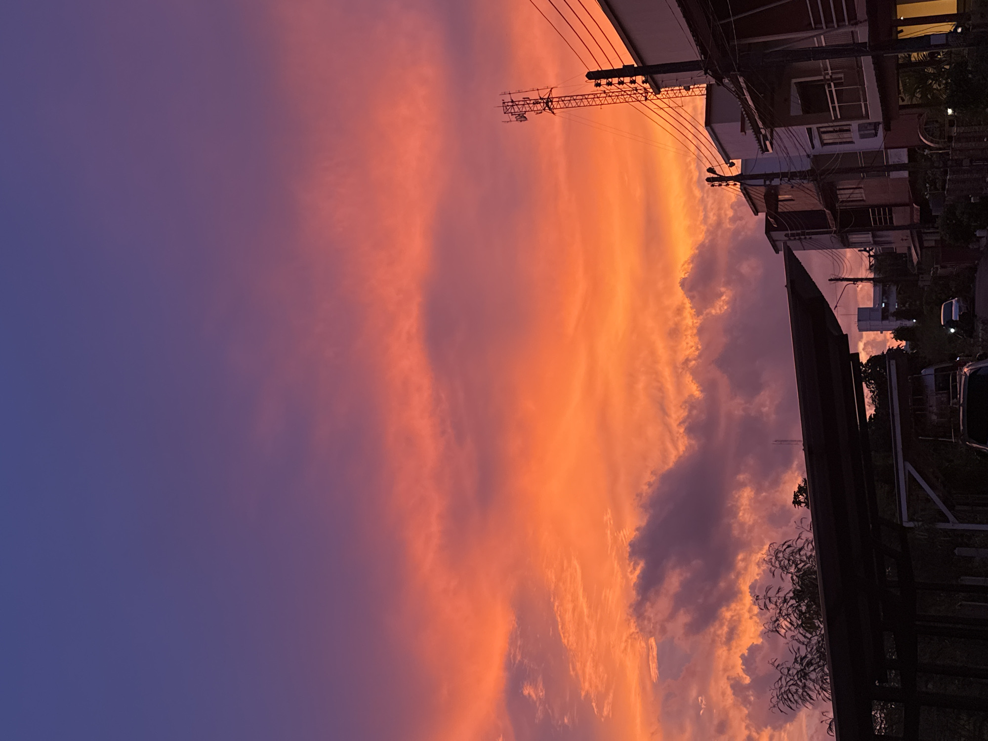 The image depicts a vibrant sunset sky over Karon, Thailand. The sky transitions from a deep purple at the top to intense shades of orange and red towards the horizon, with wispy clouds scattered throughout. In the foreground, silhouetted structures suggest buildings and possibly a carport. A cell tower is visible on the right side of the frame, along with power lines. Several multi-story buildings with balconies and windows are also present, indicating a residential or commercial area. A car is partially visible on the left, and another is seen further down a street on the left side of the image. The overall scene captures the atmospheric beauty of a sunset in an urban environment.