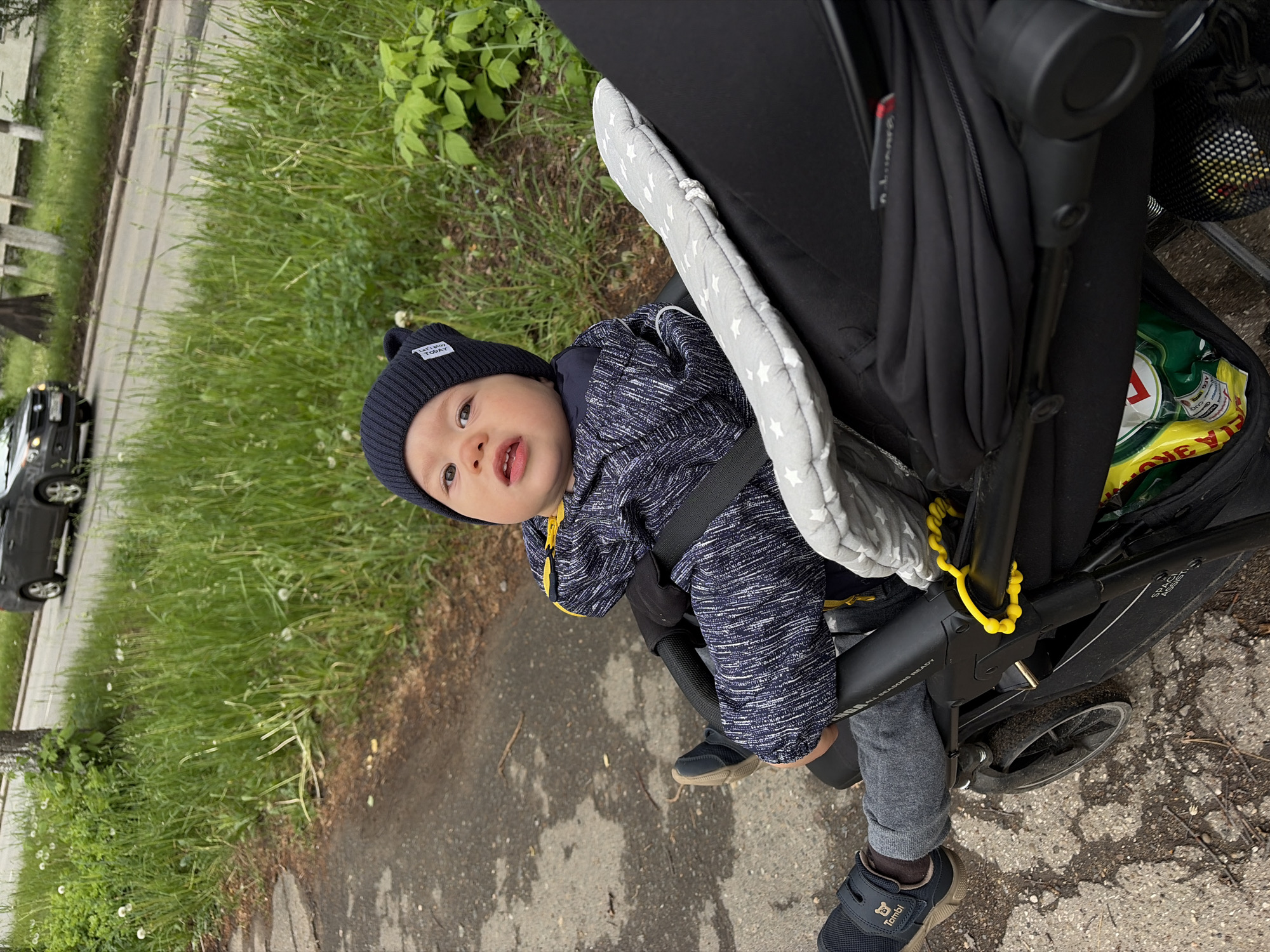 A baby is sitting in a stroller on a sidewalk beside a grassy area. A car is visible on the street in the background. The baby is wearing a blue knit hat and a blue and white jacket. A stroller umbrella is visible. A package of chips is stored in the stroller. The toddler's shoes have a bear on them. The location is likely in Chaikovsky, Russia.