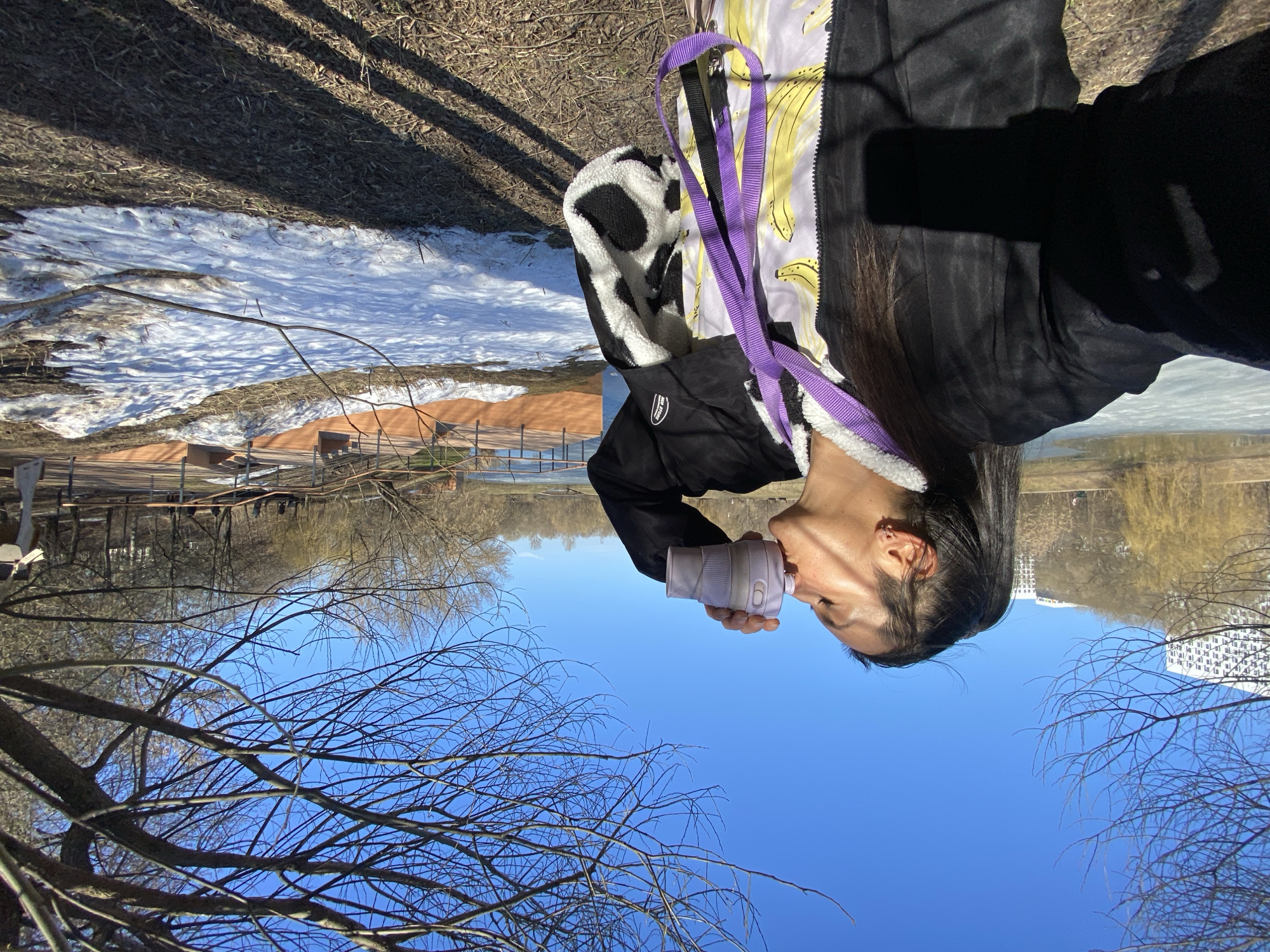 A woman is outdoors on a sunny day, drinking from a reusable water bottle. She's wearing a black jacket with a fuzzy, black-and-white cow-print lining and a banana-print bag.
The environment includes bare tree branches against a clear blue sky. In the background, there is a body of water reflecting the opposite bank, which includes snow patches, buildings, and a wooden structure. Based on the buildings in the background and the presence of a lake and trees, the location is likely a park in Moscow, Russia.