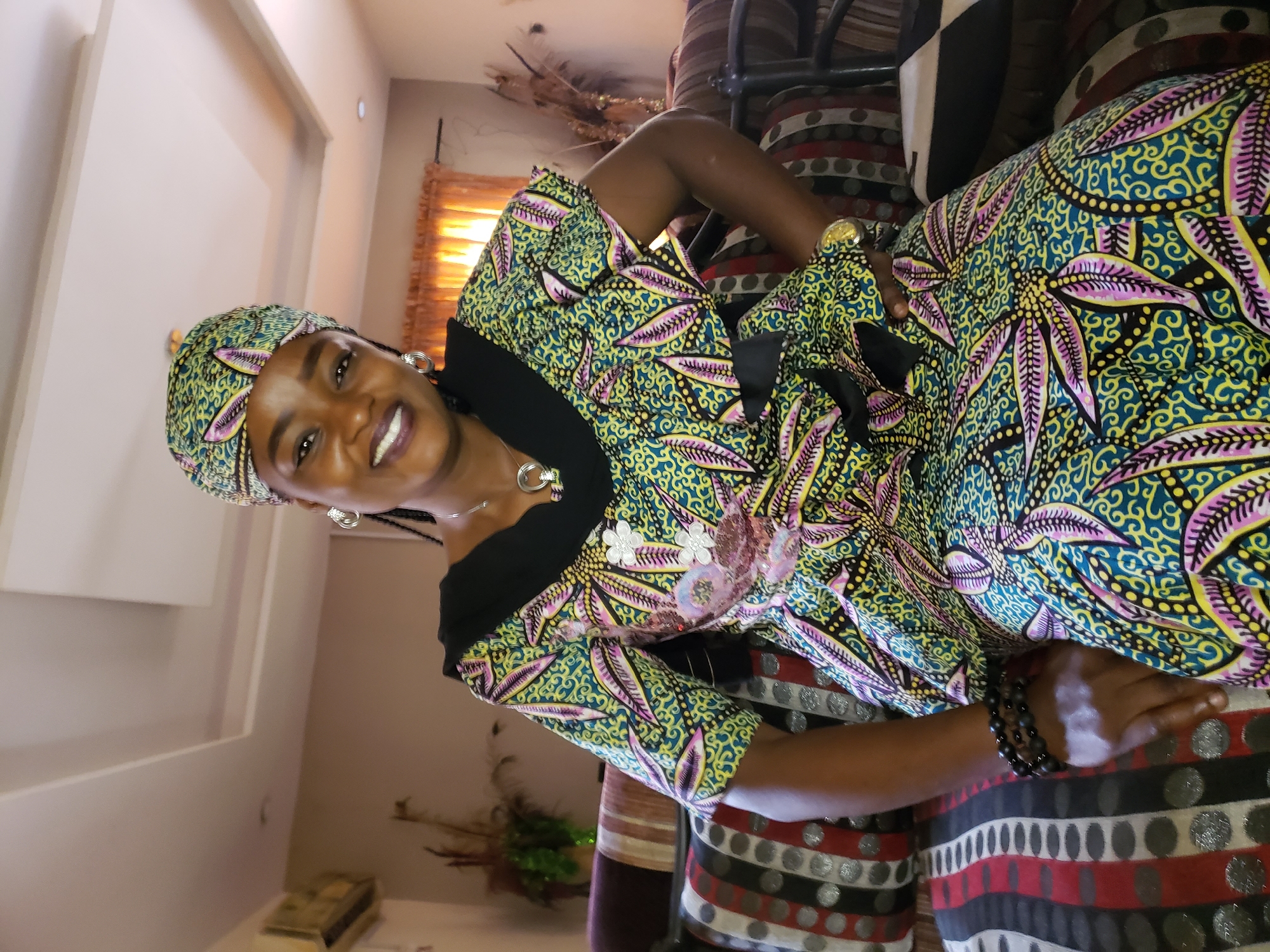 A woman with dark skin and braids, wearing a colorful headwrap and dress with a floral and geometric pattern, is seated on a sofa. The dress has a black neckline and is embellished with white floral appliques and pink sequins. She is smiling and looking towards the camera. She is wearing large hoop earrings, a silver pendant necklace, and several black beaded bracelets on her left wrist. The sofa has a striped pattern with red, white, black, and grey circles. In the background, there is an air conditioning unit on the wall, and orange curtains are visible behind her. The scene appears to be an indoor setting, possibly a living room, in Sabon Tasha, Nigeria.