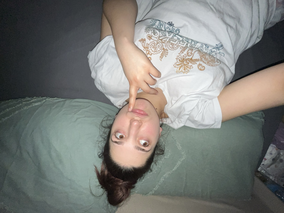 A young woman lies on a gray and green bed, looking at the camera with wide eyes. Her brown hair is tied up in a messy bun. She is wearing a white t-shirt with a printed pattern. Her left index finger is pointing towards her upper lip. The angle of the photograph is rotated, so she looks upside down.