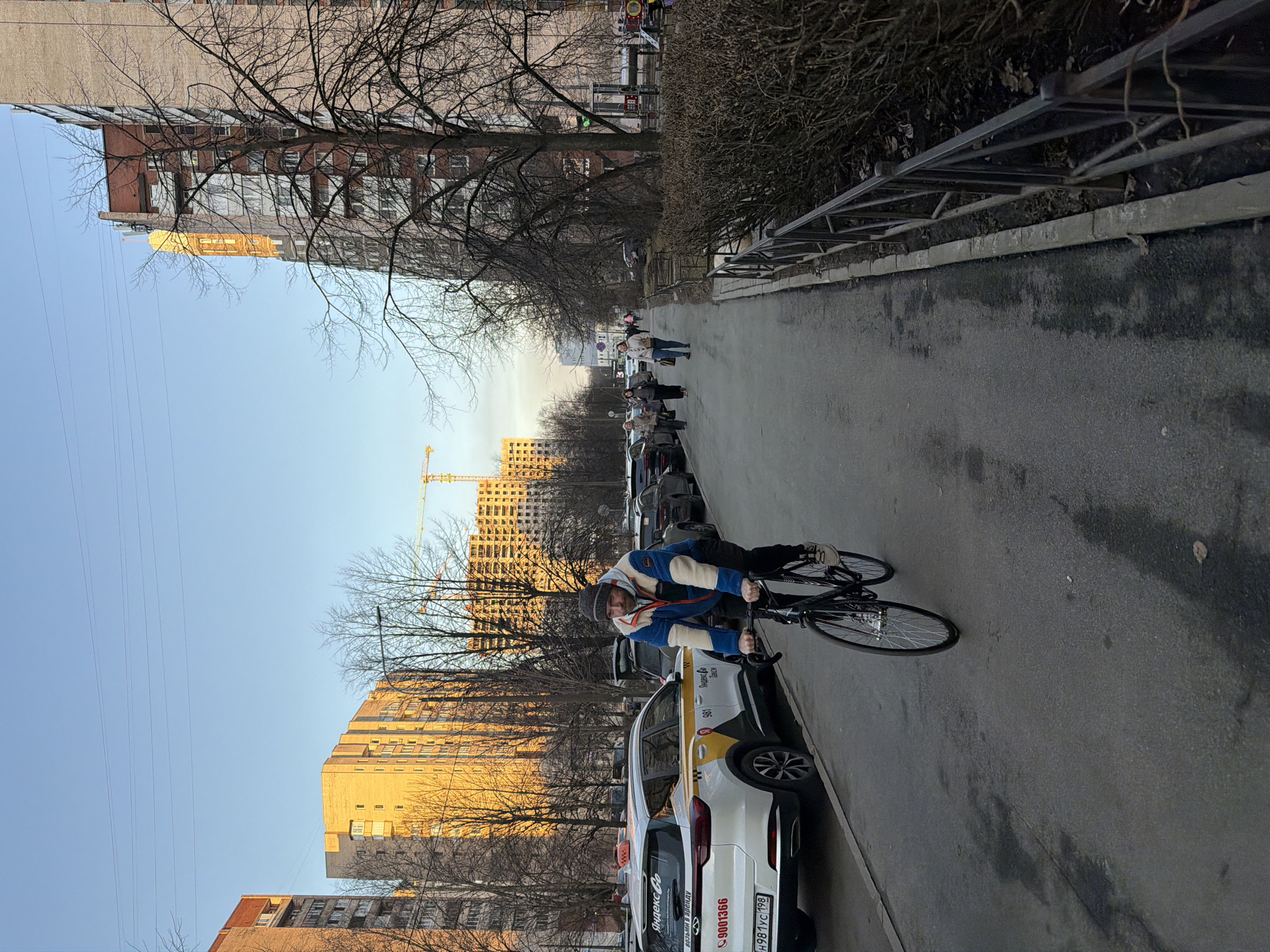 The image presents an urban street scene in Saint Petersburg, Russia, characterized by late afternoon golden hour lighting. The location is identifiable by the distinctive golden dome and cylindrical tower of the Church of the Holy Great Martyr George the Victorious (located at Engels Prospekt, 111k1), visible in the background.
In the immediate foreground, a bearded male cyclist, appearing to be middle-aged, rides a black road bicycle on an asphalt road. He wears a grey knit beanie, a blue, white, and red fleecy jacket, dark pants, and light-colored shoes, and is looking towards the camera. To the cyclist's left, a line of parked vehicles includes a prominent white Yandex Go taxi, a Chery Tiggo 4 Pro, featuring yellow and grey branding and Russian license plate H981YC 198 (Saint Petersburg region code 198). Further along the street, additional dark-colored cars are parked.
To the cyclist's right, a paved sidewalk accommodates several pedestrians walking away from the camera, including two women in the mid-ground. The sidewalk is separated from a dense row of bare bushes by a low, dark metal fence. Beyond the bushes and fence, bare deciduous trees are interspersed among multi-story residential buildings. These buildings, constructed of brick and concrete, are illuminated by the low-angle sunlight, creating strong contrasts. In the far background, a yellow construction crane indicates ongoing development against a clear, light blue sky traversed by faint power lines. The overall environment suggests either late autumn, winter, or early spring due to the lack of foliage on trees and bushes.