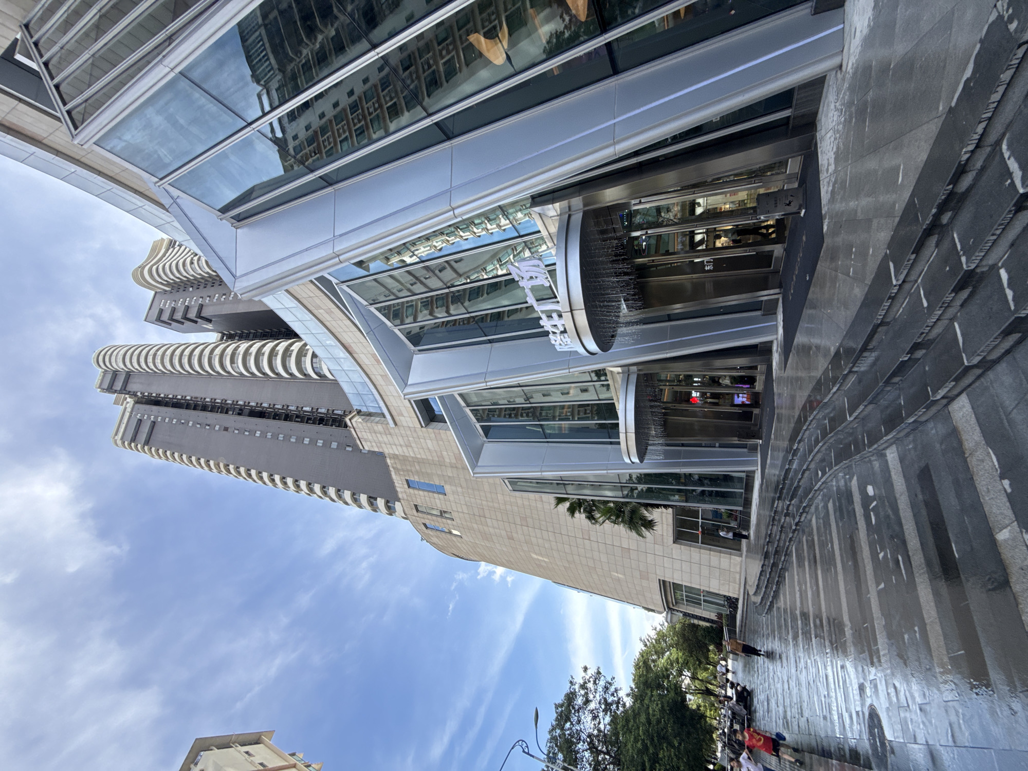 The image depicts the exterior of the Longsheng Plaza, a modern commercial building in Huizhou, China. The building features a distinctive, angular architectural design with extensive use of glass and metallic elements. In the foreground, a wet, stone-paved walkway with tiered steps curves away from the entrance, suggesting recent rain. Several individuals are visible on the walkway, with some walking and others appearing to be children running. To the left of the plaza, trees line the street, and a street lamp is present. In the background, several high-rise residential buildings with curved balconies dominate the skyline. The sky is partly cloudy. The signage on the building's entrance appears to be in Chinese characters.