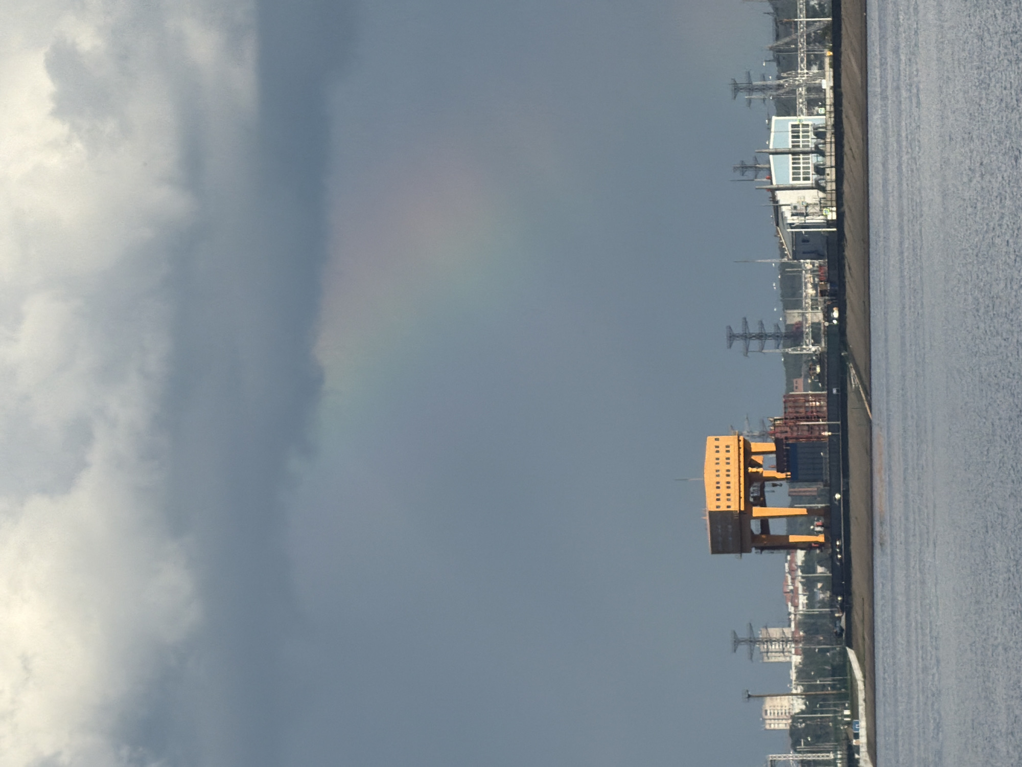 The image presents a panoramic view of a waterfront industrial area, likely a port, under a dramatic sky. The top portion of the frame is dominated by a cloudy sky, featuring a blend of bright white and dark gray cumulus clouds. A faint, ethereal rainbow arc is subtly visible in the upper central sky, suggesting recent rain or atmospheric moisture.
Below the sky, the left side of the frame showcases a developed cityscape along the water's edge. A prominent, large orange or yellow gantry-like structure, possibly a massive crane or part of a shipyard, stands out in the mid-ground. Various other industrial buildings and structures, including a light blue building with multiple white windows, dot the landscape. Numerous tall, skeletal structures, resembling power line towers or port equipment, are scattered throughout, indicating a bustling industrial zone. A dark, linear strip runs along the immediate waterfront, appearing to be a road, pier, or quay.
The right side of the image is filled by a wide expanse of water, which appears calm but with subtle surface texture, reflecting the overhead light.
No specific human activity is discernible, and no people are visible. The scene captures the interplay between large-scale industrial infrastructure and natural atmospheric phenomena. The time of day is daytime, likely during or after a rain shower, given the presence of the rainbow and clouds. The setting is a developed waterfront, consistent with a port or heavy industrial area, potentially in Noviy, Russia. No visible text is present.